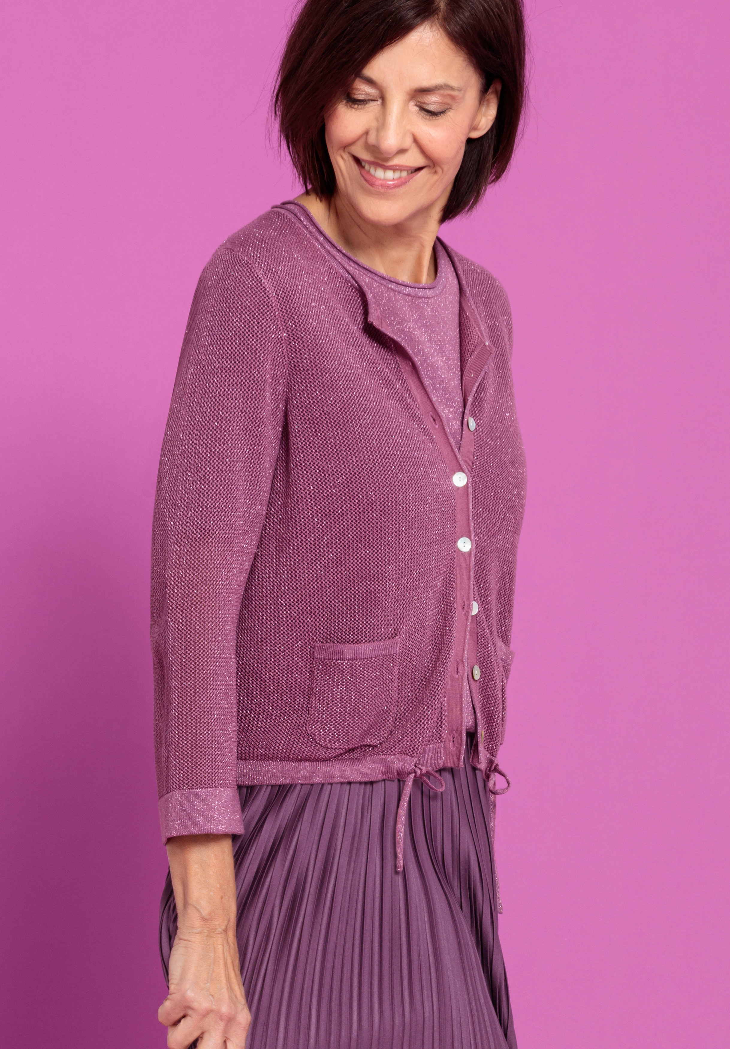 Eine Frau mit kurzen braunen Haaren lächelt und blickt nach unten. Sie trägt eine malvenfarbene Strickjacke mit weißen Knöpfen über einem passenden Oberteil und einem Faltenrock und steht vor einem hellrosa Hintergrund.