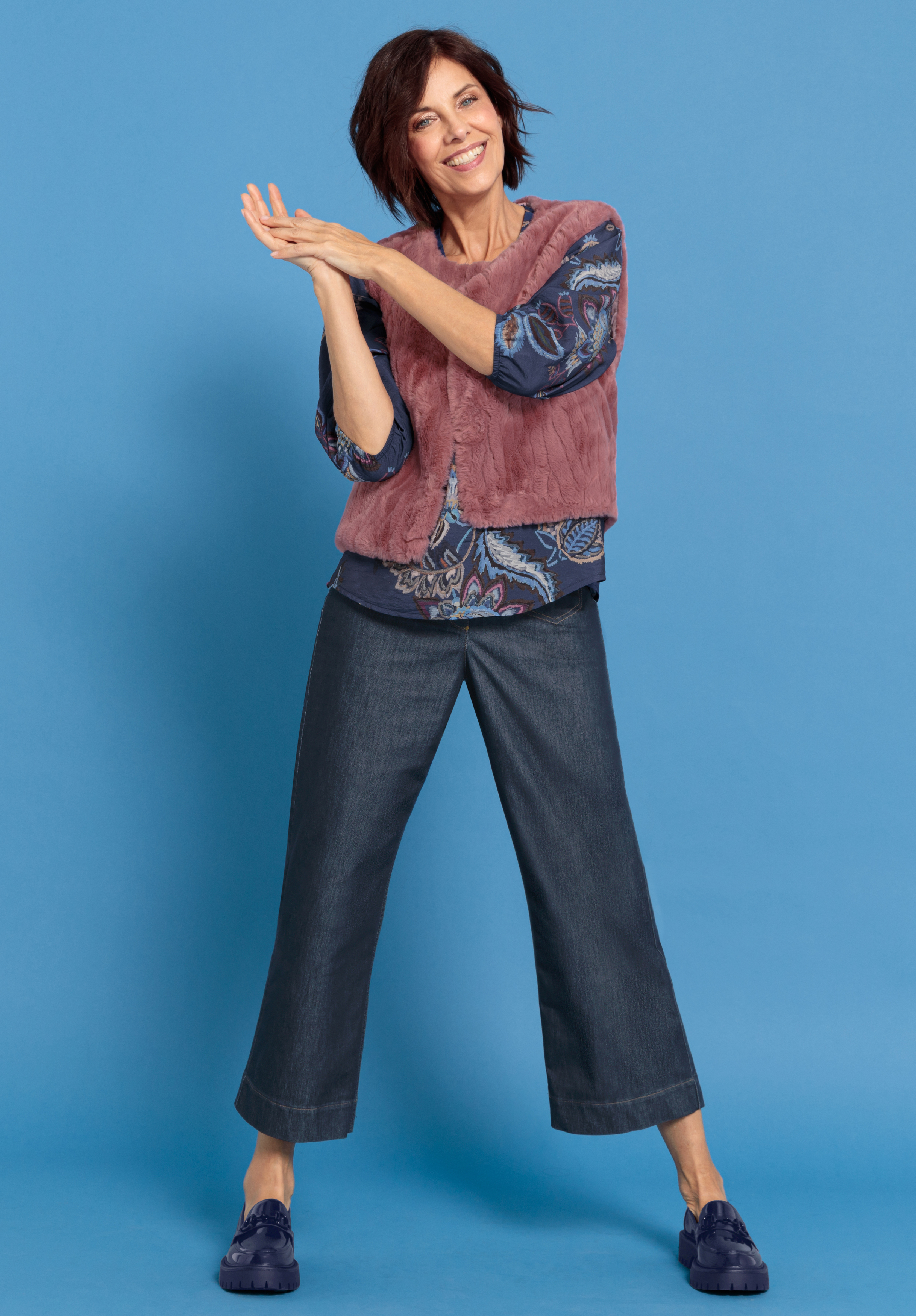 Eine lächelnde Frau mit kurzen dunklen Haaren steht vor einem blauen Hintergrund. Sie trägt eine lilafarbene Weste über einem gemusterten Oberteil, dunkle Jeans mit weitem Bein und marineblaue Schuhe. Sie klatscht in einer fröhlichen Pose in die Hände.