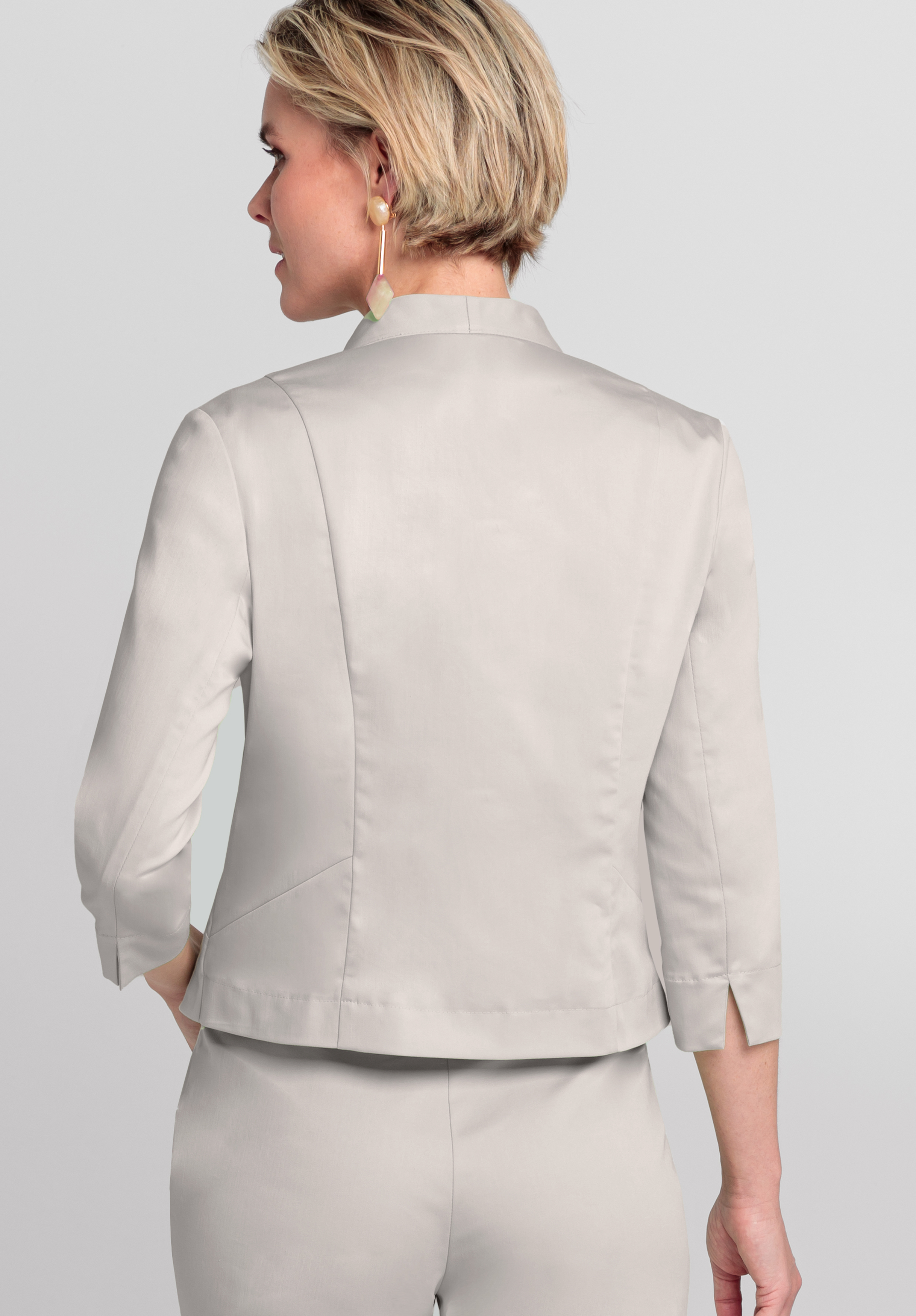 Eine Frau mit kurzen blonden Haaren, Ohrhängern und einem hellgrauen Blazer mit Dreiviertelärmeln und Rückennähten steht mit dem Rücken zur Kamera. Zum Blazer trägt sie eine passende Hose.