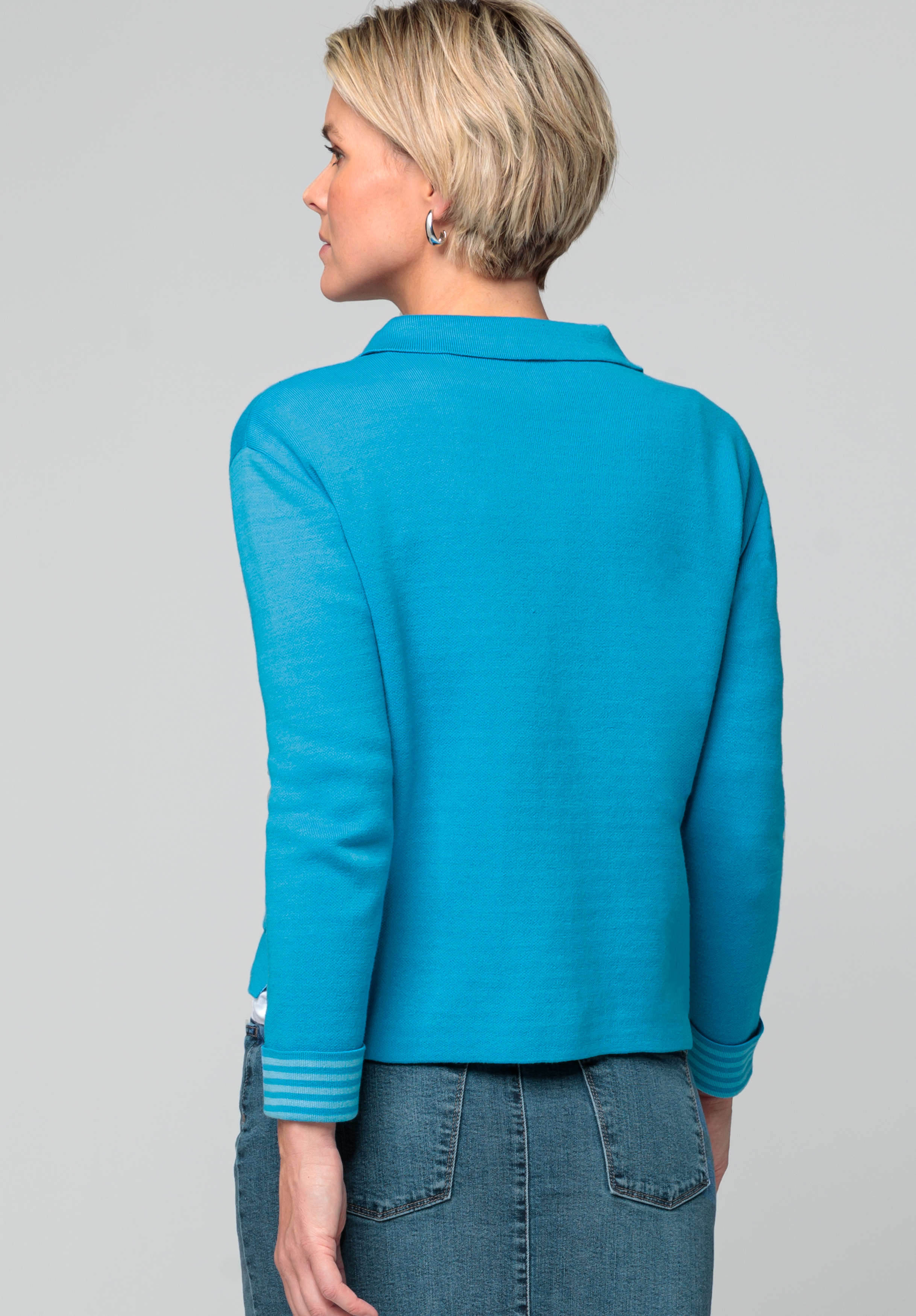 Eine Frau mit kurzen blonden Haaren, von hinten gesehen, trägt eine leuchtend blaue Langarmjacke und blaue Jeans. Sie steht vor einem schlichten hellgrauen Hintergrund.