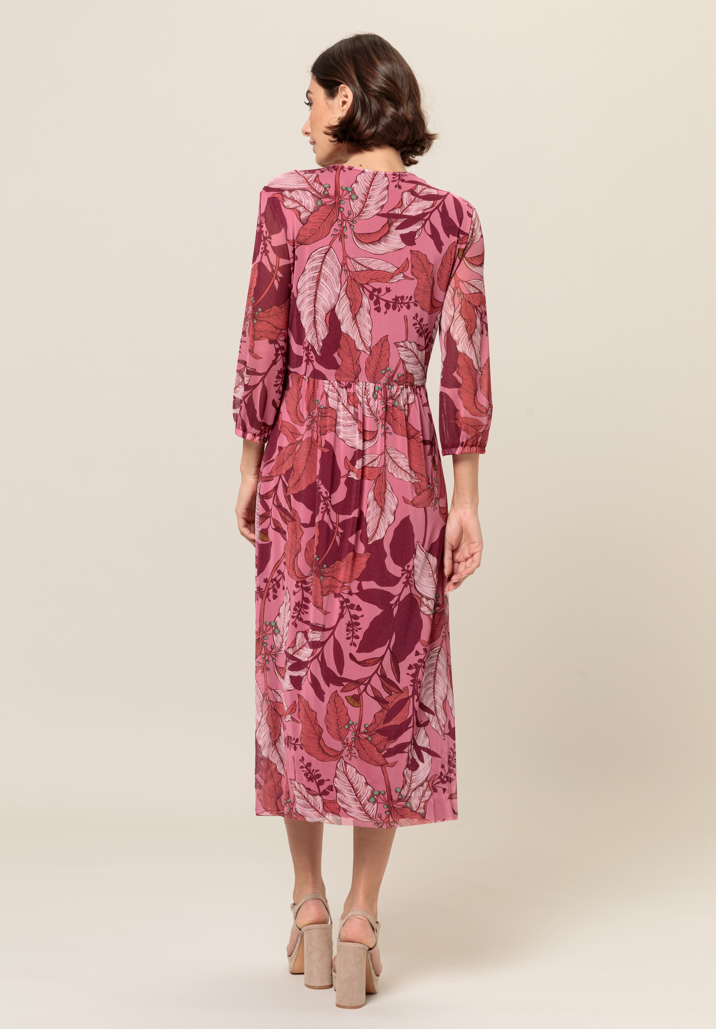 Eine Frau steht mit abgewandtem Gesicht da. Sie trägt ein langärmeliges Midikleid mit rosa-kastanienbraunem Blattmuster. Sie hat kurzes braunes Haar und trägt beige Sandaletten vor einem schlichten Hintergrund.