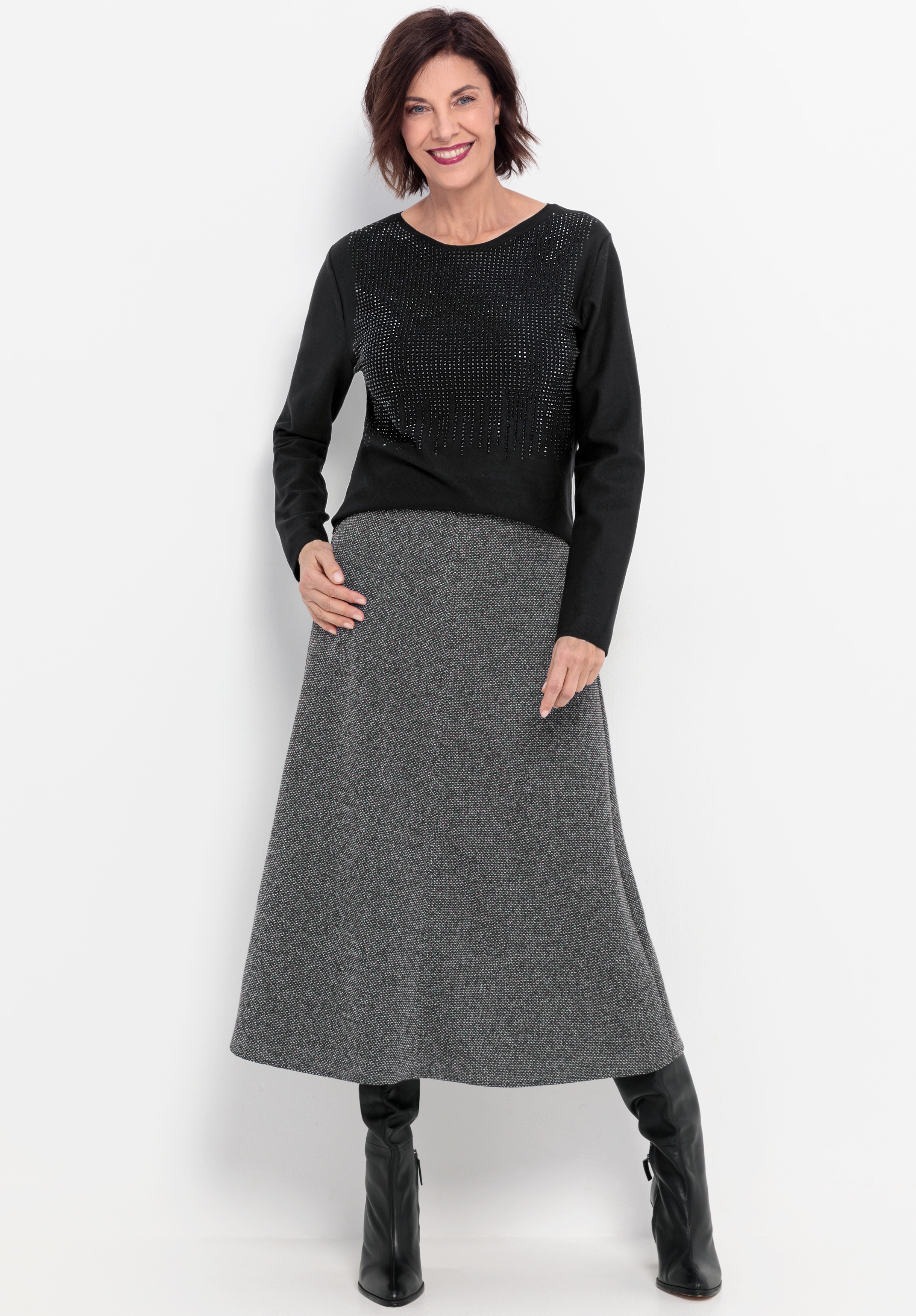 Eine Frau steht lächelnd vor einem weißen Hintergrund und trägt ein schwarzes Langarm-Oberteil mit dezenten Verzierungen, einen grauen Midirock in A-Linie und schwarze kniehohe Stiefel.