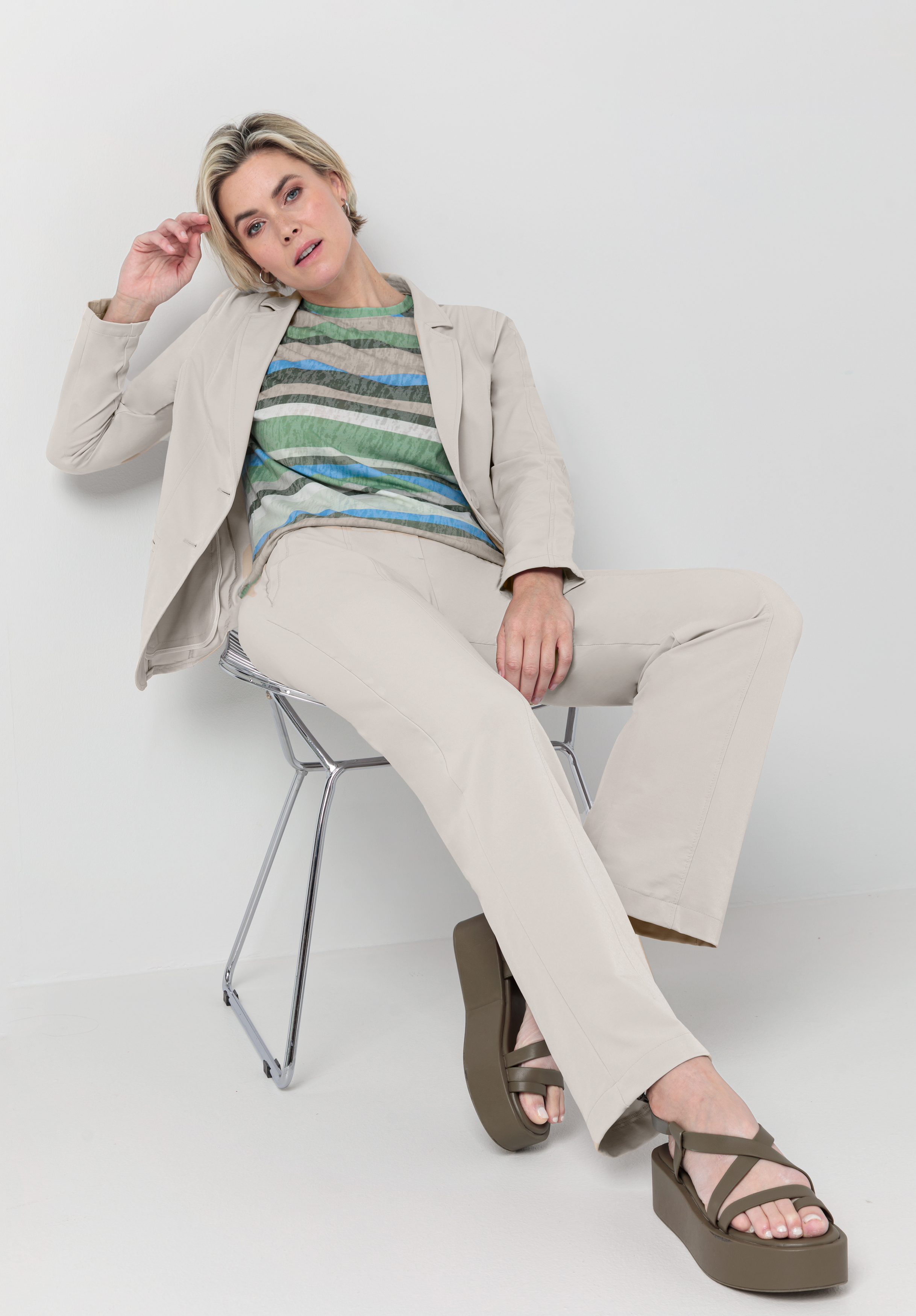Eine Person mit kurzen blonden Haaren sitzt lässig auf einem modernen Stuhl. Sie trägt einen hellbeigen Anzug, ein bunt gestreiftes Oberteil und braune Plateausandalen vor weißem Hintergrund.