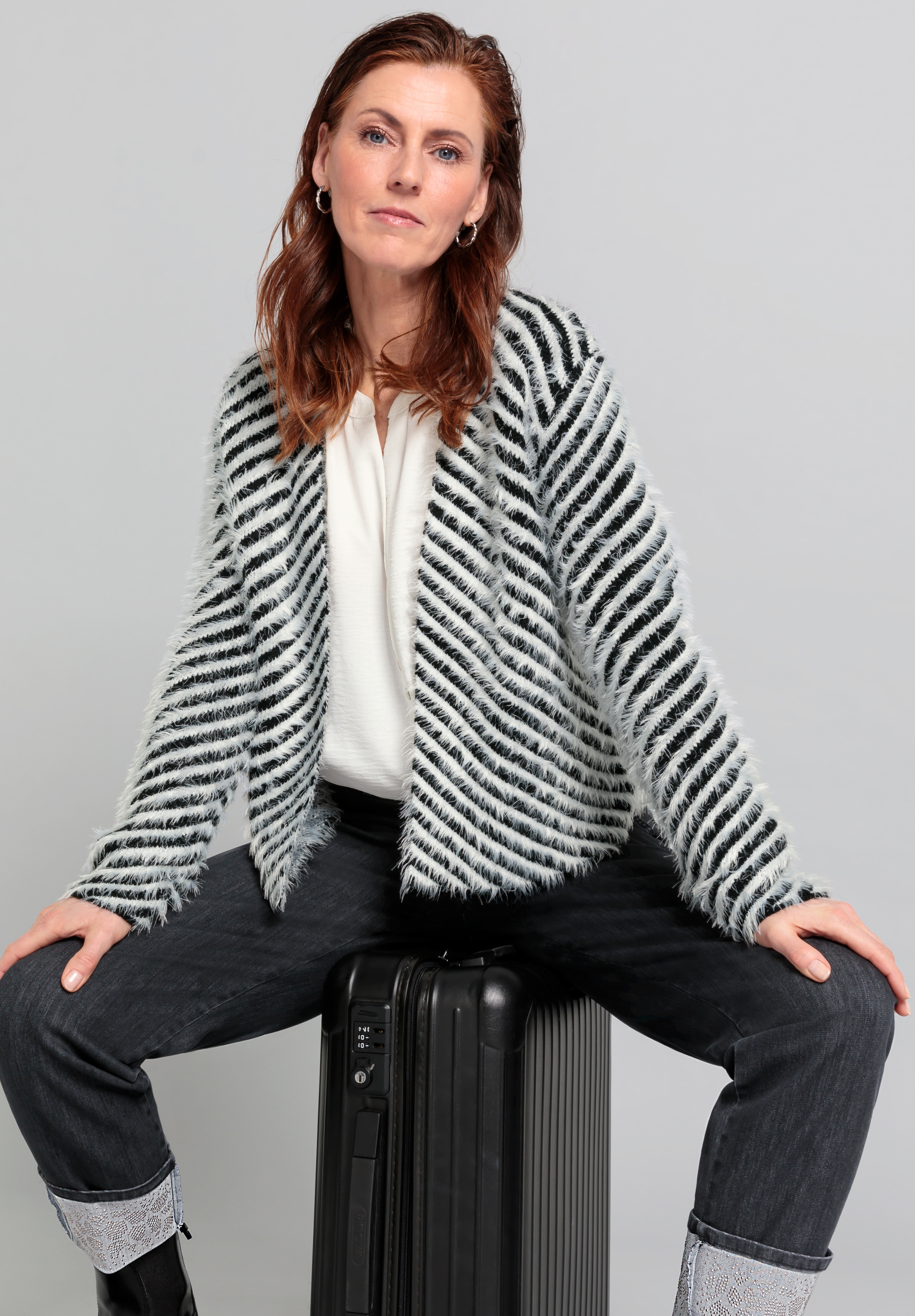 Eine Frau mit rotbraunem Haar, die eine gestreifte, flauschige Jacke, ein weißes Oberteil und eine dunkle Jeans trägt, sitzt selbstbewusst auf einem schwarzen Koffer vor einem schlichten grauen Hintergrund.