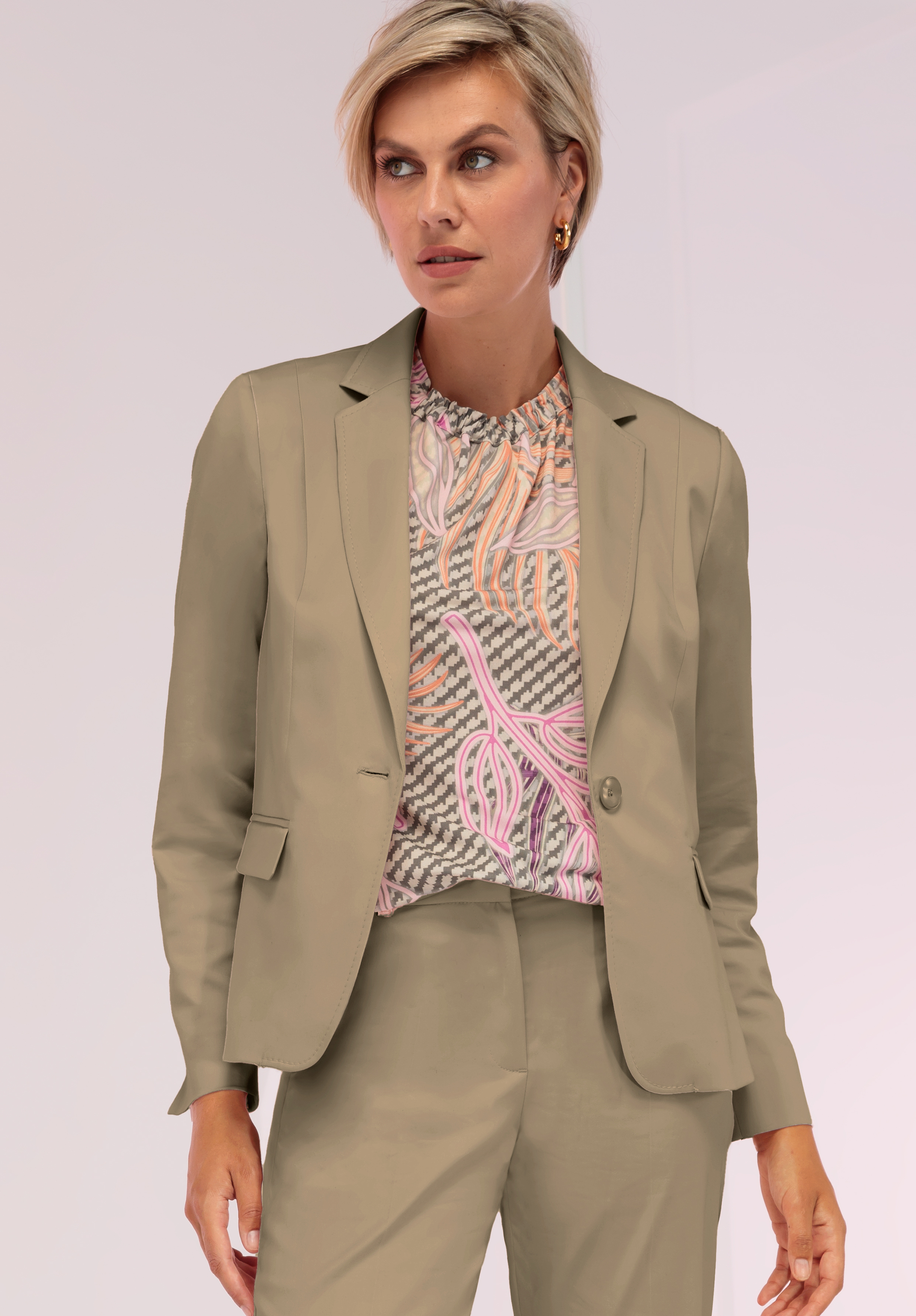 Eine Frau mit kurzen blonden Haaren trägt ein hell olivgrünes Kostüm mit einem Einknopf-Blazer und passender Hose, dazu eine bunt gemusterte Bluse. Ihr Blick ist vor einem hellen Hintergrund leicht zur Seite gerichtet.