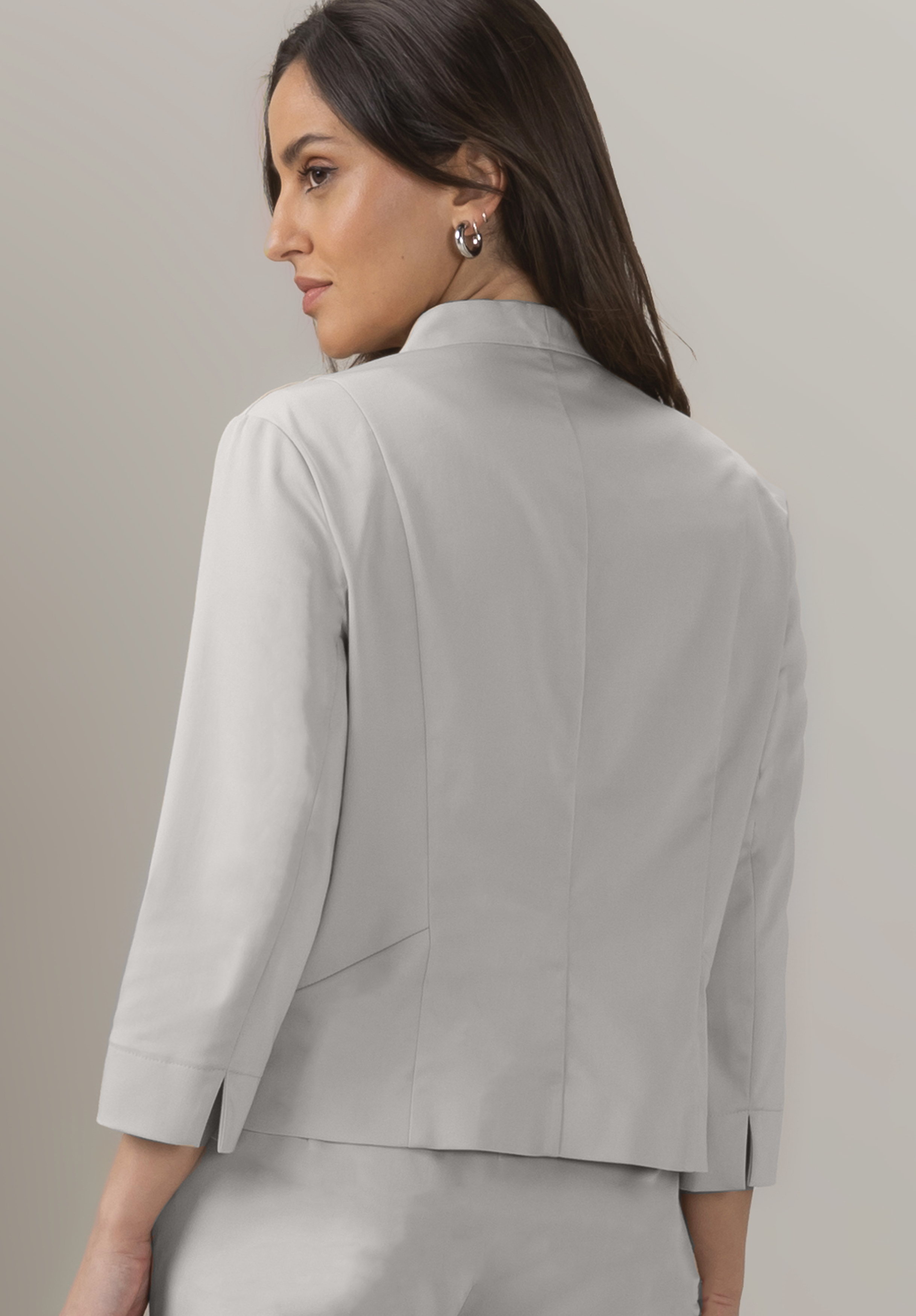 Eine Frau mit langen dunklen Haaren, einem hellgrauen Blazer und einer dazu passenden Hose, ist von hinten zu sehen und dreht den Kopf leicht zur Seite. Der Hintergrund ist schlicht und neutral.