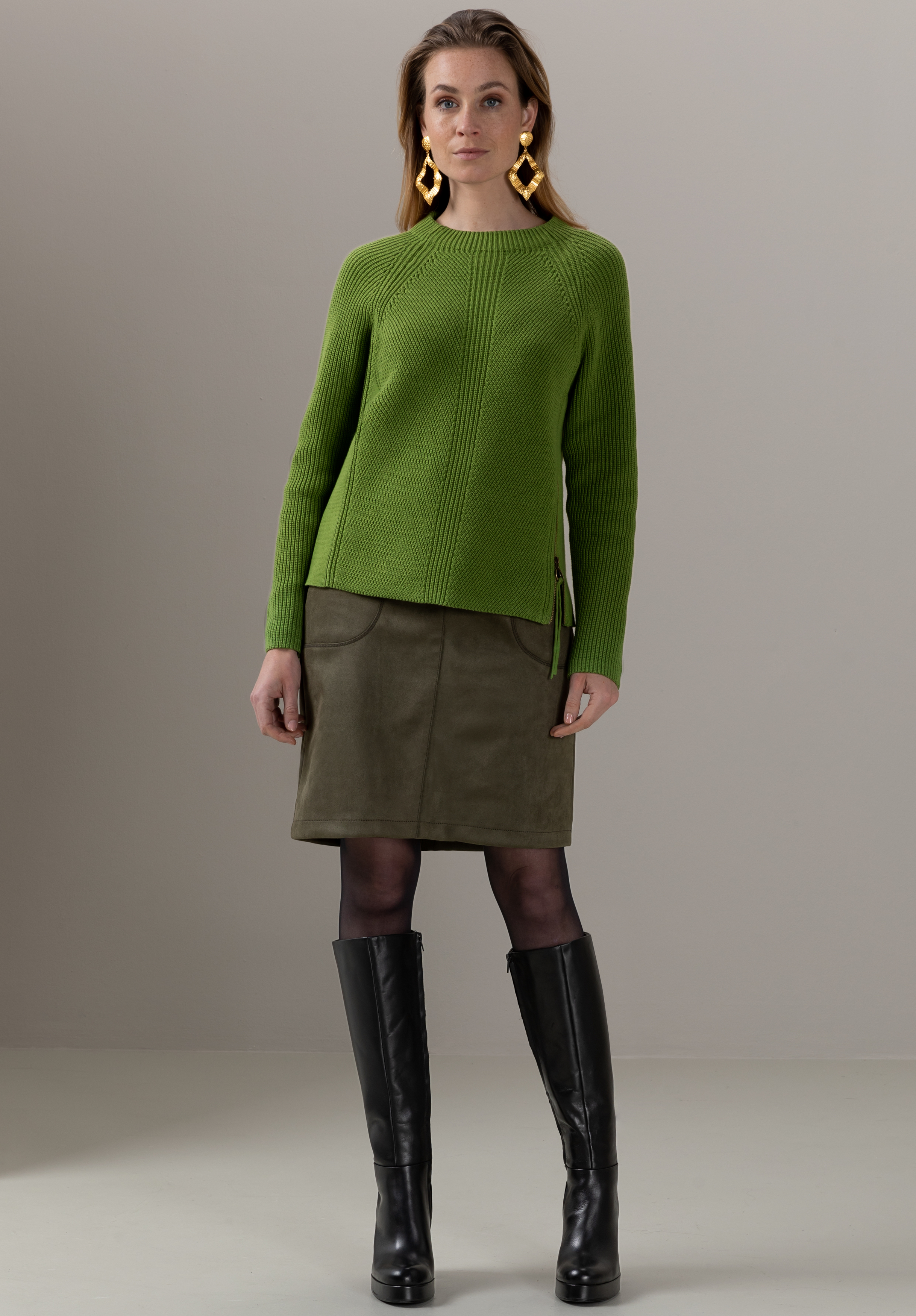 Eine Frau in einem grünen Strickpullover, einem olivgrünen Rock, schwarzen Strumpfhosen, kniehohen schwarzen Stiefeln und großen goldenen Ohrringen steht vor einem schlichten beigen Hintergrund.