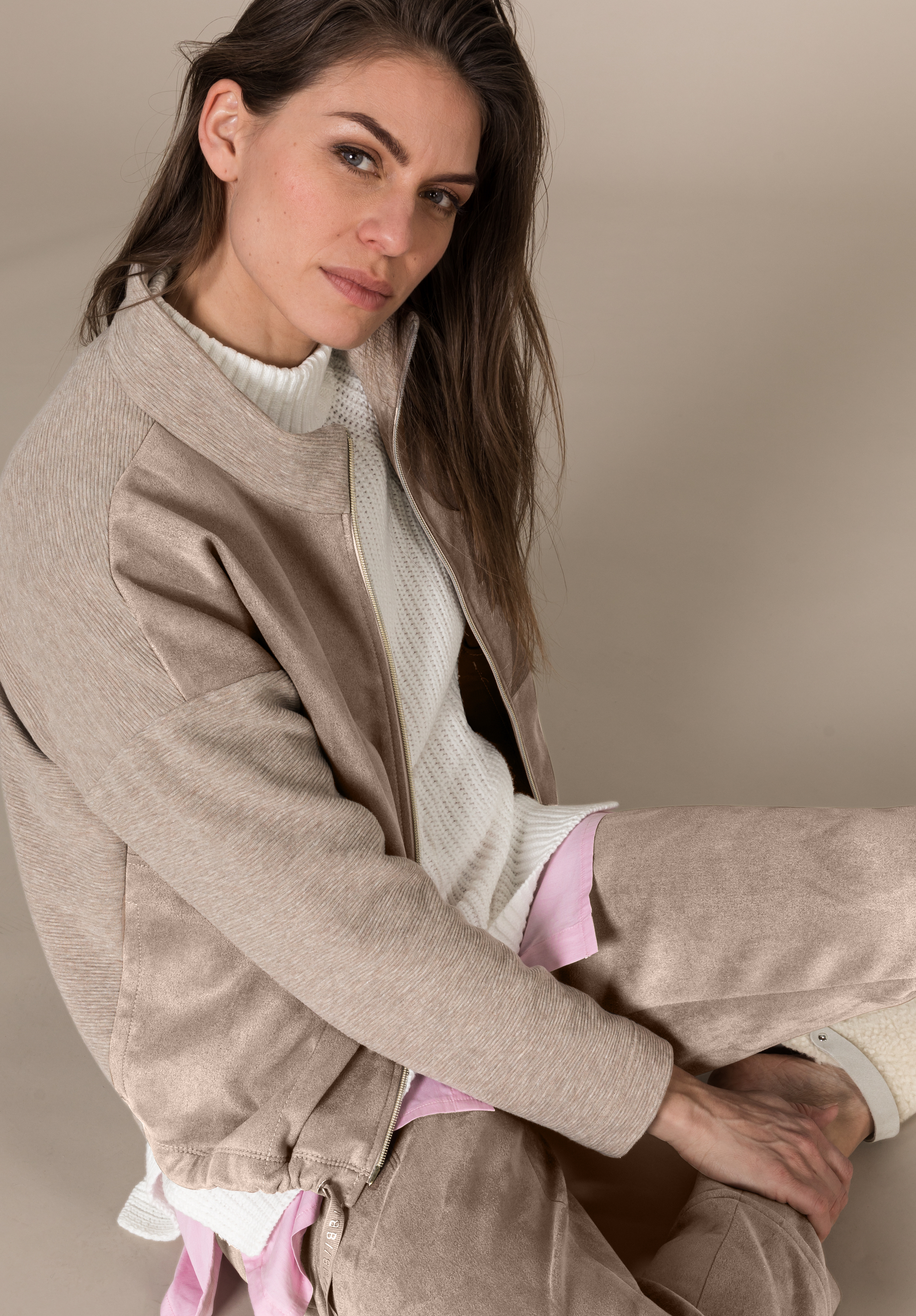 Eine Frau mit langen braunen Haaren, die eine beige Jacke, einen weißen gerippten Rollkragenpullover und eine dazu passende beige Hose trägt, sitzt vor einem neutralen Hintergrund auf dem Boden und blickt leicht nach oben in die Kamera.