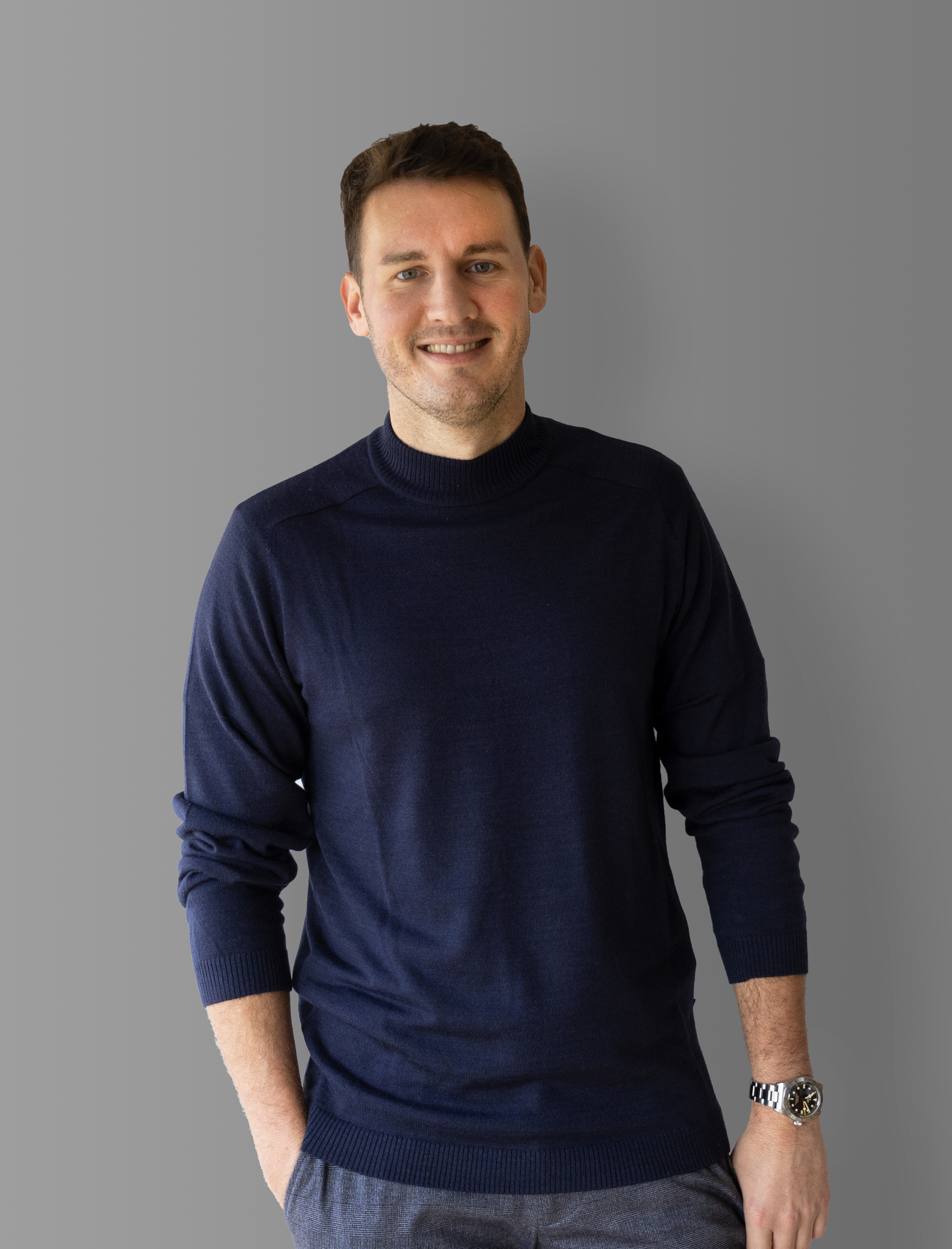 Ein Mann mit kurzen braunen Haaren, der einen marineblauen Pullover und eine graue Hose trägt, steht lächelnd vor einem schlichten grauen Hintergrund und hat die Hände in den Taschen.