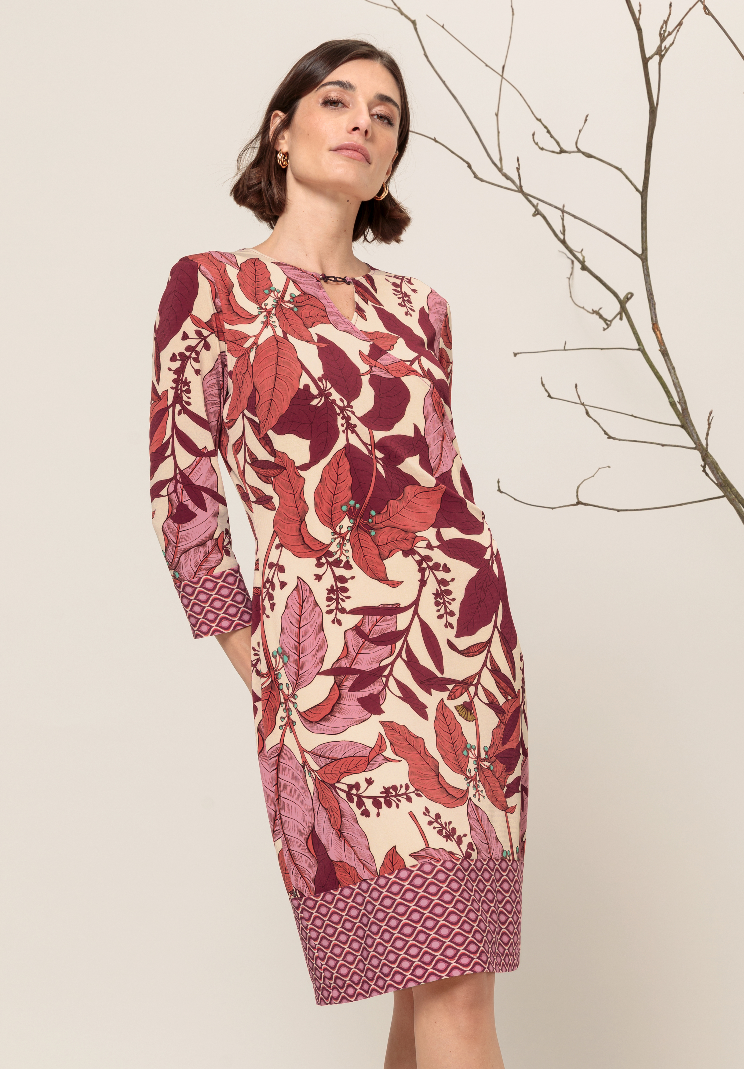 Eine Frau in einem beigen Kleid mit kräftigen roten und rosafarbenen botanischen Drucken steht selbstbewusst neben einem kahlen Ast vor einem neutralen Hintergrund.