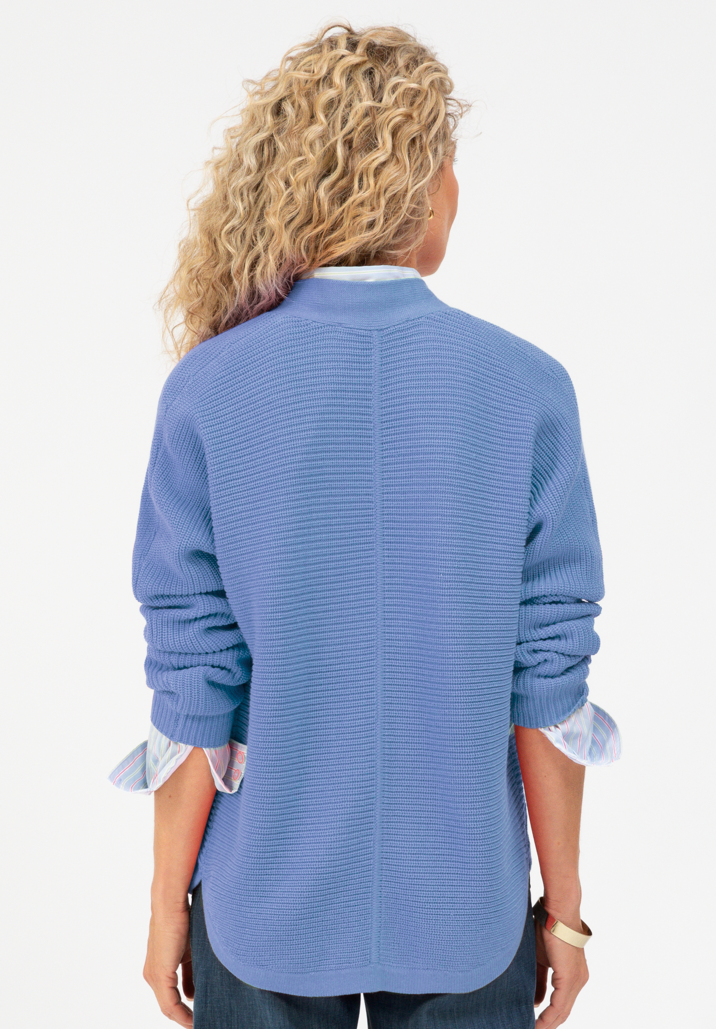 Eine Person mit lockigem blondem Haar, von hinten gesehen, trägt einen strukturierten blauen Pullover über einem gestreiften Hemd mit hochgekrempelten Ärmeln sowie eine dunkle Hose. Der Hintergrund ist schlicht weiß.