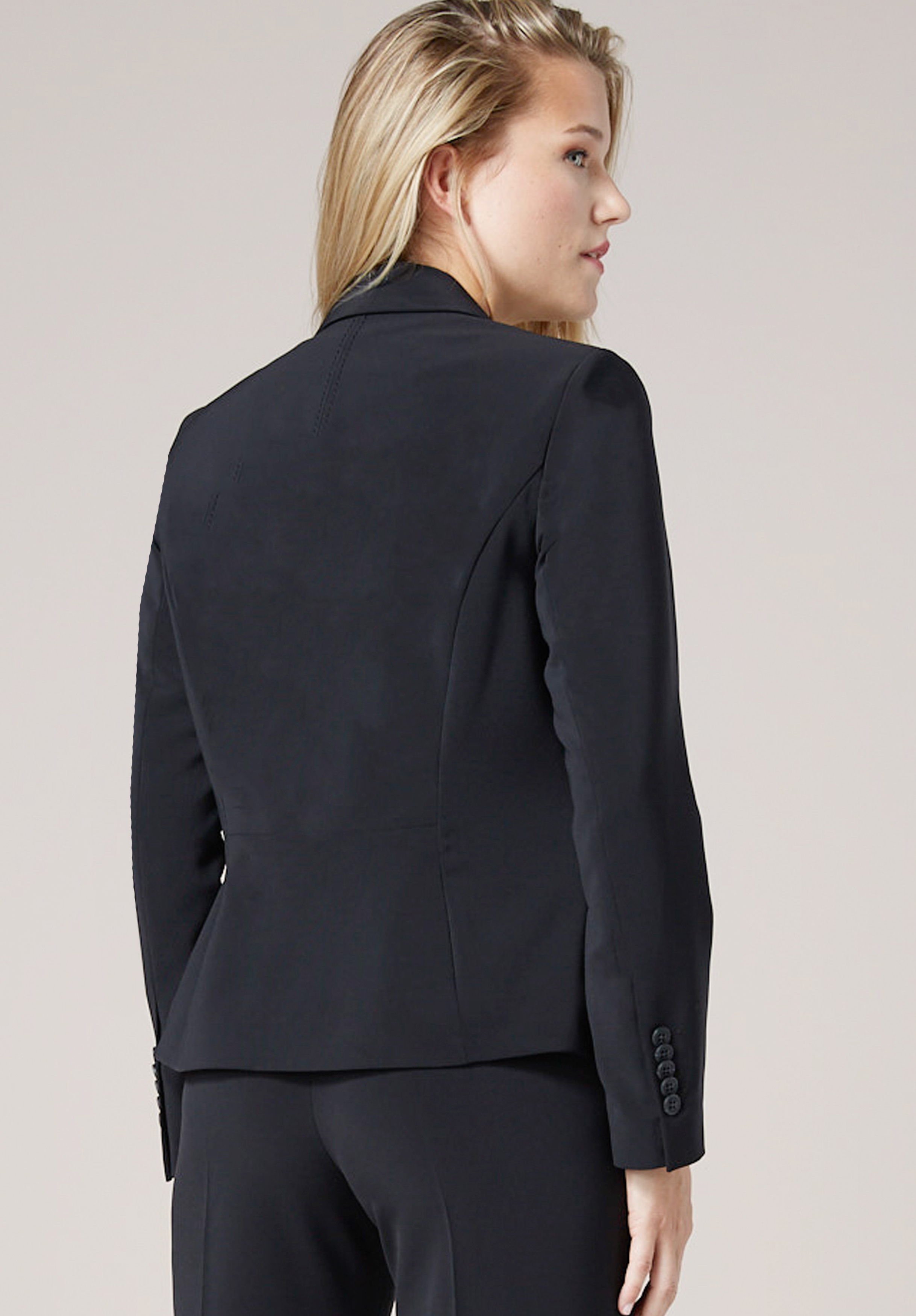 Eine Frau mit blondem Haar, die einen taillierten schwarzen Blazer und eine dazu passende Hose trägt, steht mit abgewandtem Gesicht da und blickt vor einem schlichten hellen Hintergrund zur Seite.