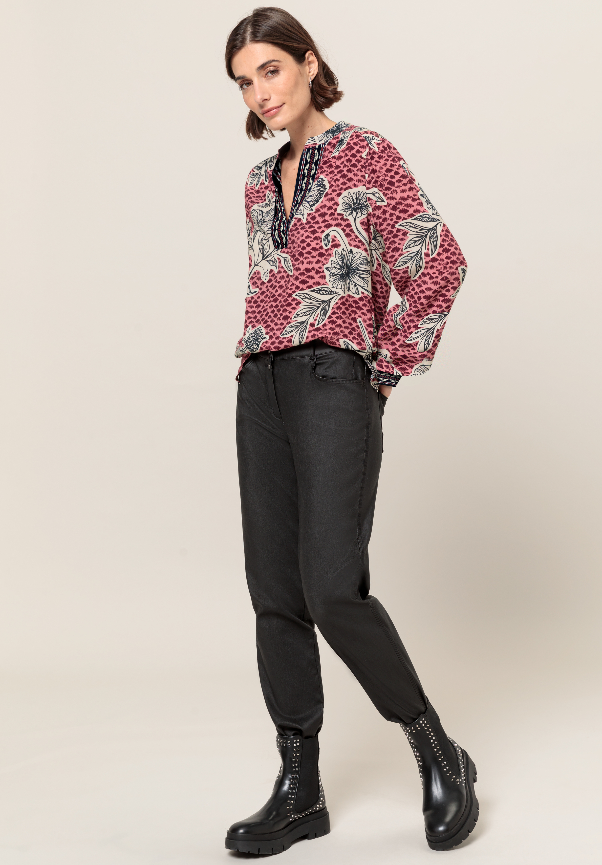Eine Frau steht vor einem schlichten Hintergrund. Sie trägt eine rote Bluse mit Blumenmuster, eine schwarze Hose mit hoher Taille und schwarze Stiefeletten mit Nieten.