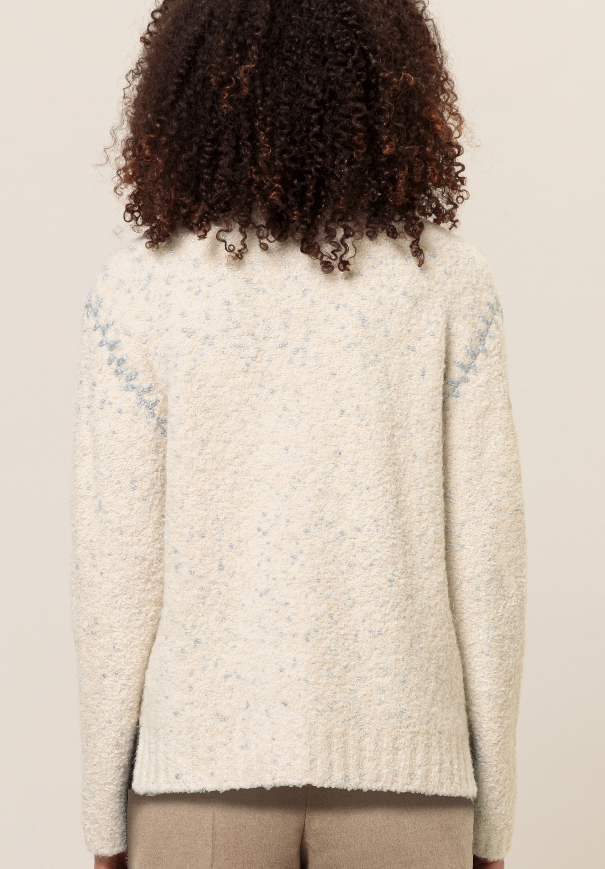 Von hinten ist eine Person mit lockigem Haar zu sehen, die einen strukturierten, cremefarbenen Pullover mit dezenten blauen Sprenkeln und beige Hosen vor einem schlichten Hintergrund trägt.