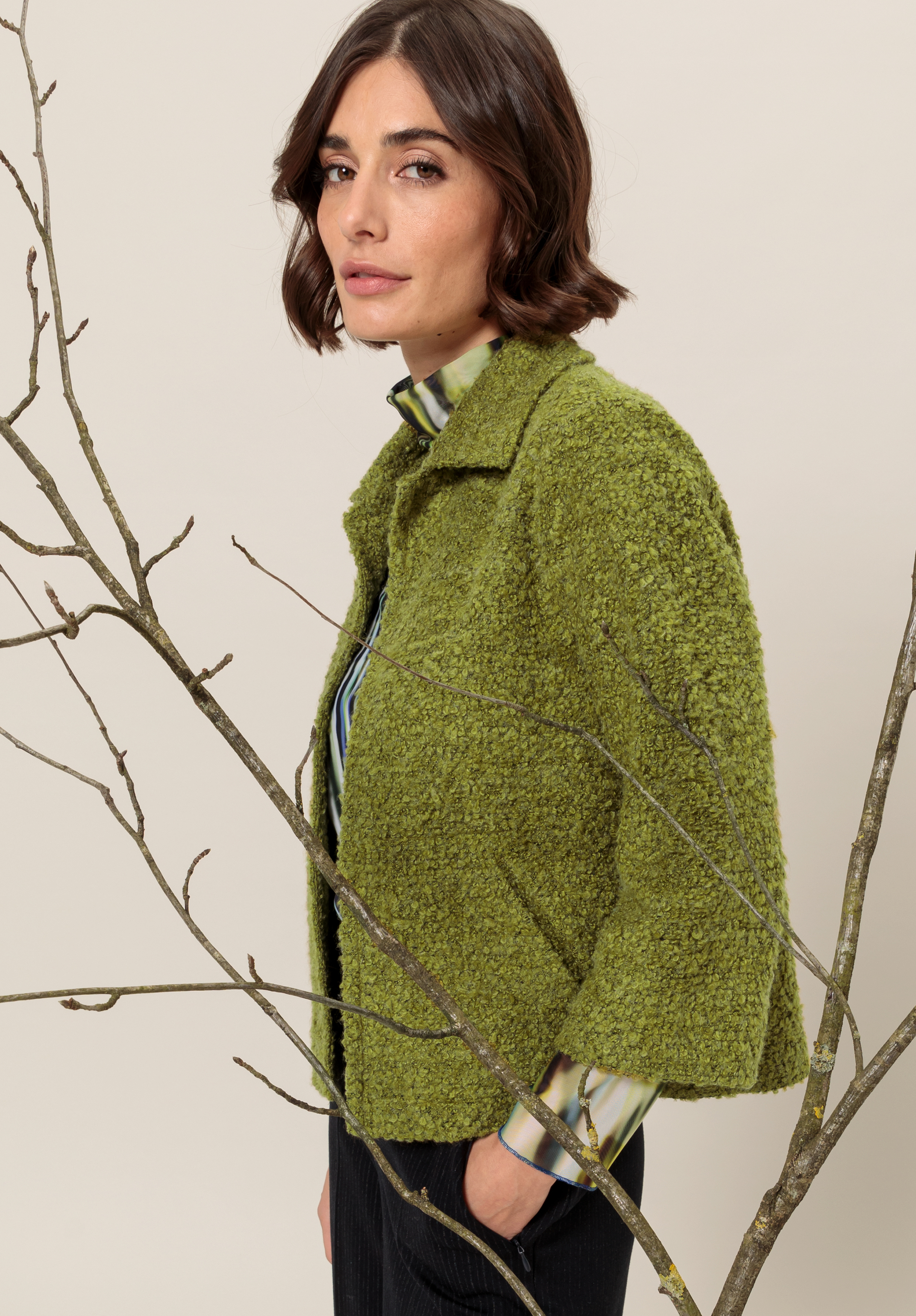 Eine Frau mit kurzen braunen Haaren trägt eine strukturierte grüne Jacke über einem karierten Hemd und steht seitlich neben kahlen Ästen vor einem neutralen Hintergrund.