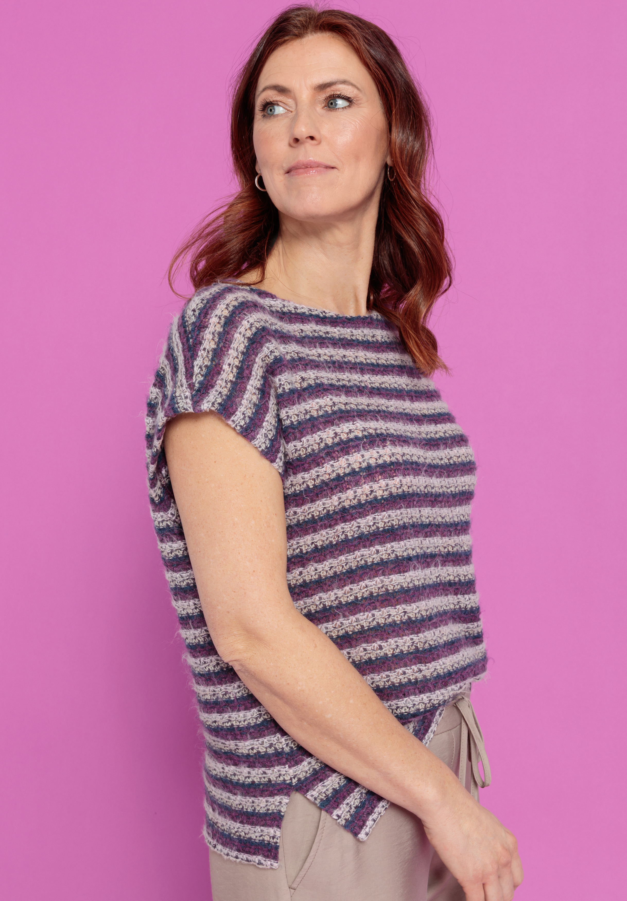 Eine Frau mit gewelltem kastanienbraunem Haar trägt ein kurzärmeliges, lila und grau gestreiftes Strickoberteil und eine beigefarbene Hose. Sie steht vor einem einfarbig rosafarbenen Hintergrund und blickt leicht zur Seite.