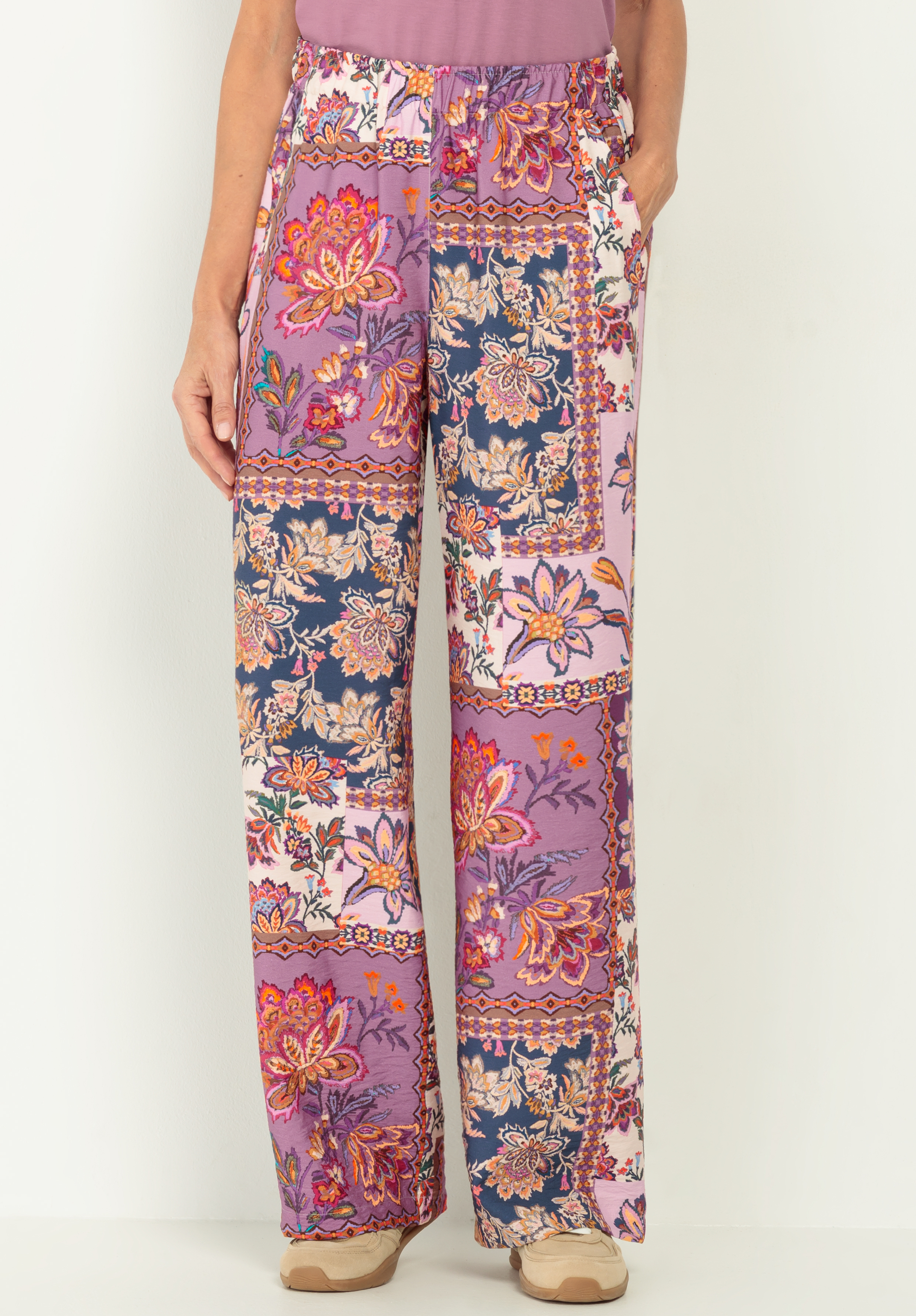 Eine Person trägt eine lockere Hose mit weitem Bein und einem bunten Patchwork-Blumenmuster in Lila-, Rosa-, Marine- und Orangetönen. Die Hose hat Seitentaschen und einen elastischen Bund. Nur der Unterkörper ist sichtbar.