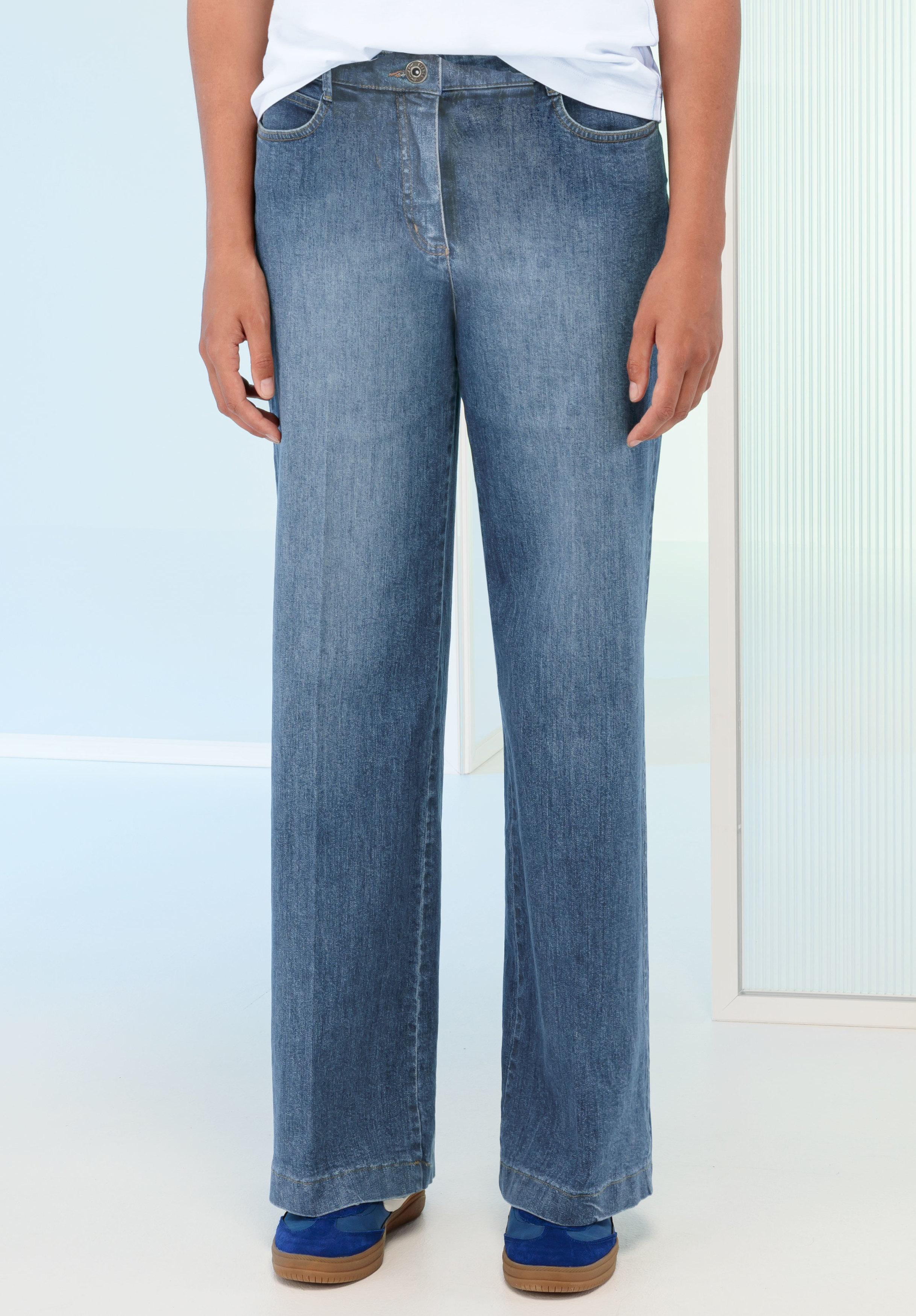 Eine Person, die eine weit geschnittene Bluejeans und blaue Schuhe trägt, steht in einem Innenraum vor einem hellen Hintergrund. Ihre Hände liegen entspannt an der Seite und ihr Oberkörper ist teilweise sichtbar.