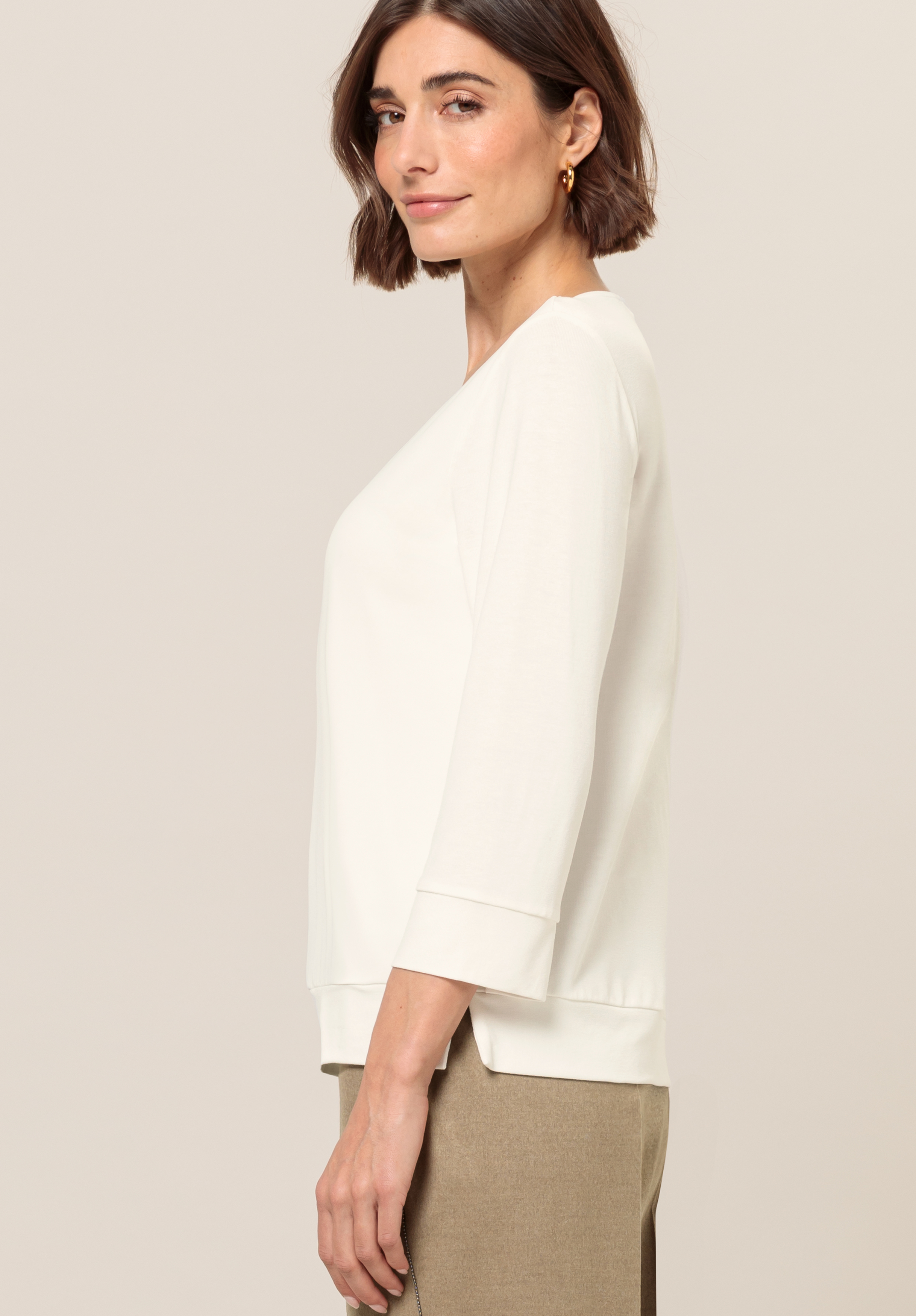 Eine Frau mit kurzen braunen Haaren, die ein weißes Oberteil mit Dreiviertelärmeln und eine beige Hose trägt, steht im Profil vor einem schlichten, hellen Hintergrund.