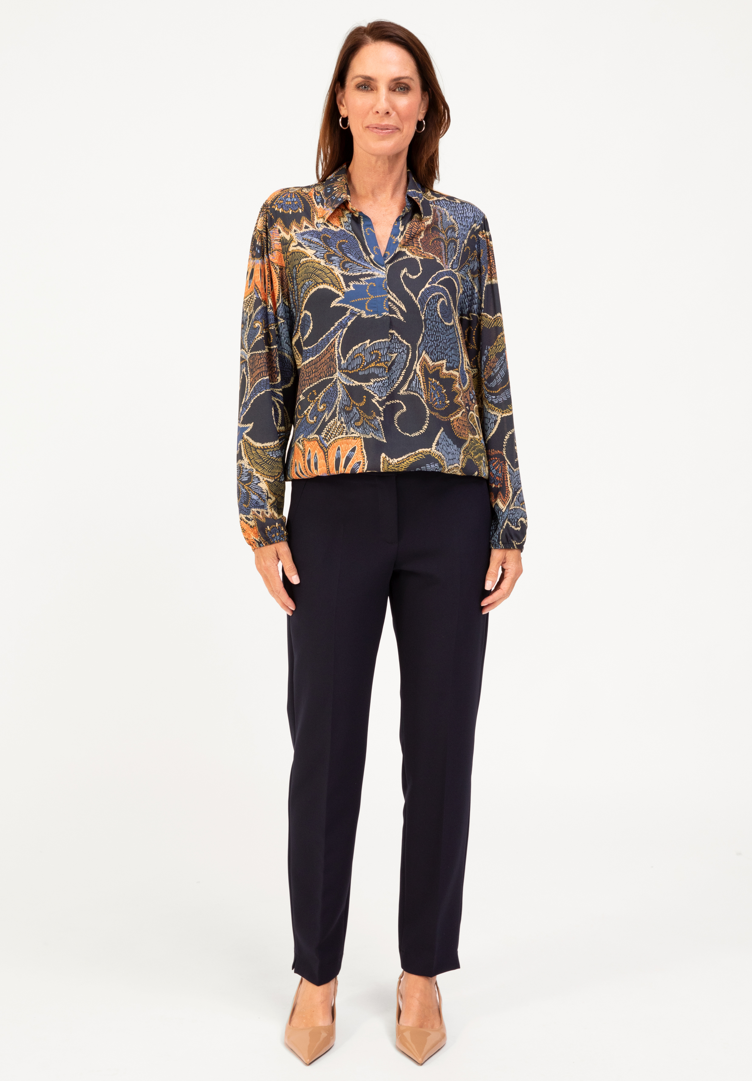 Eine Frau steht mit dem Gesicht nach vorne, trägt eine langärmelige, bunt gemusterte Bluse, eine marineblaue Hose und beigefarbene spitze Absätze vor einem schlichten weißen Hintergrund.