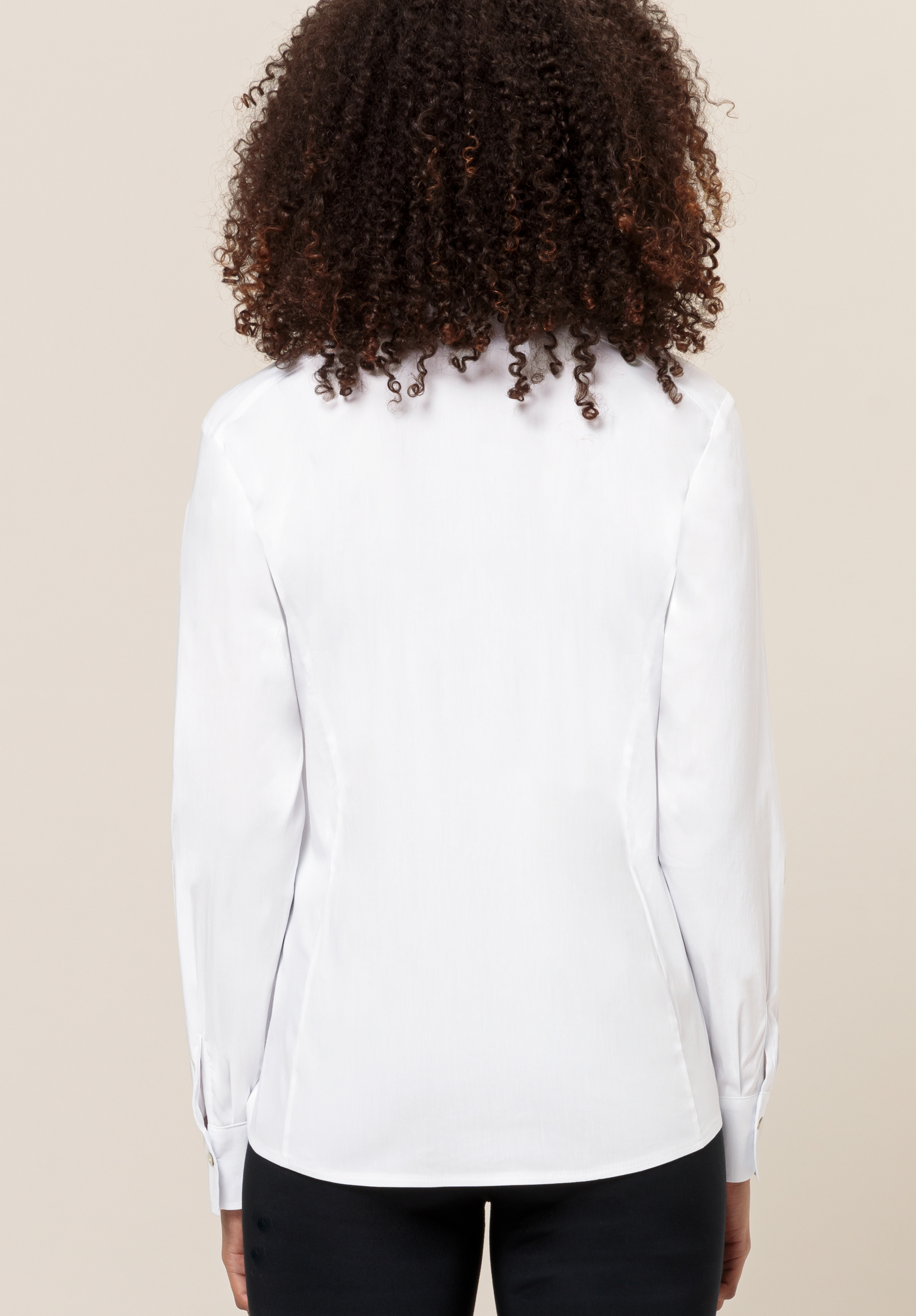 Gezeigt wird eine Person mit lockigem Haar von hinten, die eine langärmelige weiße Bluse und eine schwarze Hose trägt und vor einem schlichten beigen Hintergrund steht.
