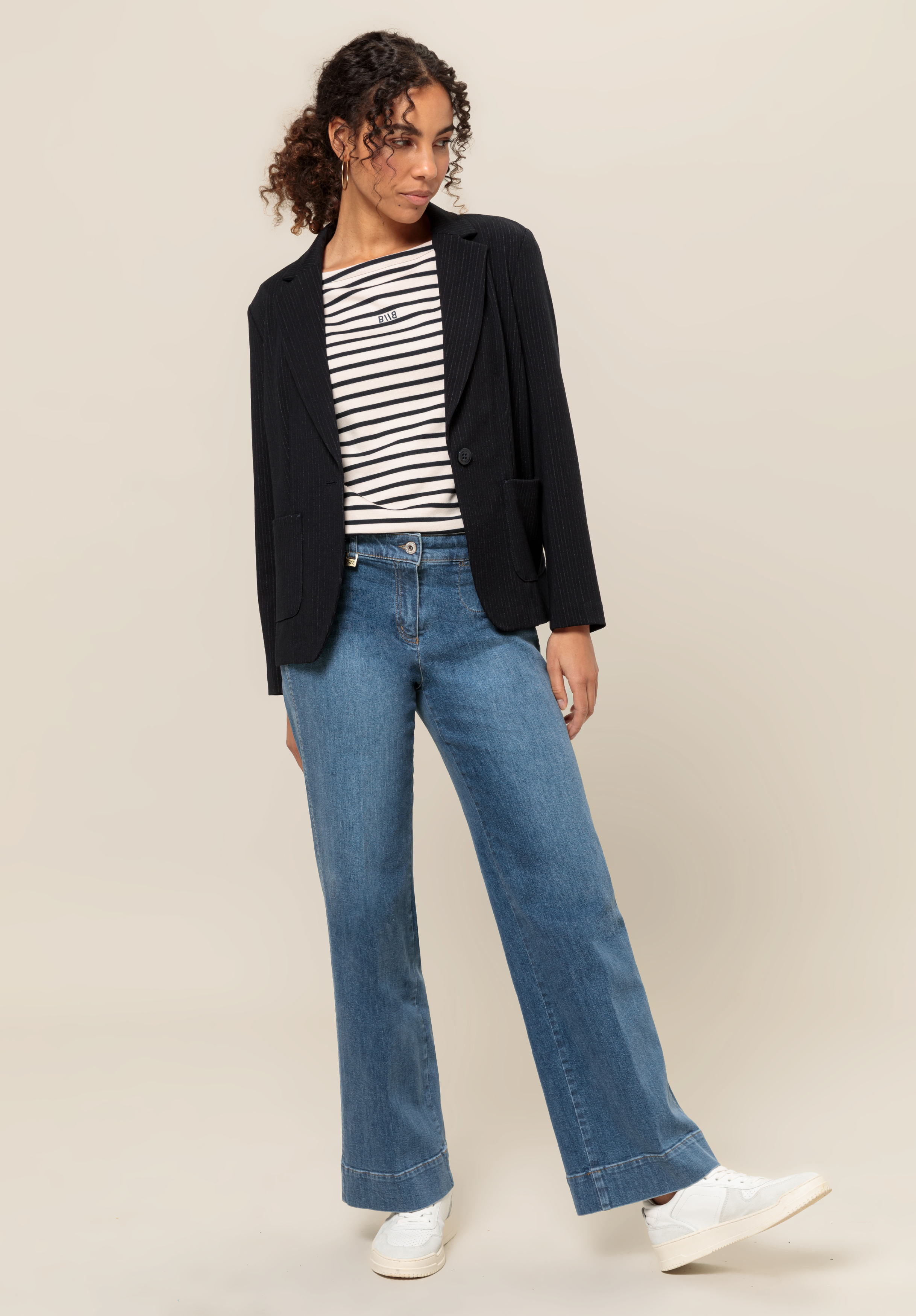 Eine Frau mit lockigem Haar trägt einen schwarzen Blazer über einem schwarz-weiß gestreiften Hemd, weite Bluejeans und weiße Turnschuhe und steht vor einem schlichten hellbeigen Hintergrund.