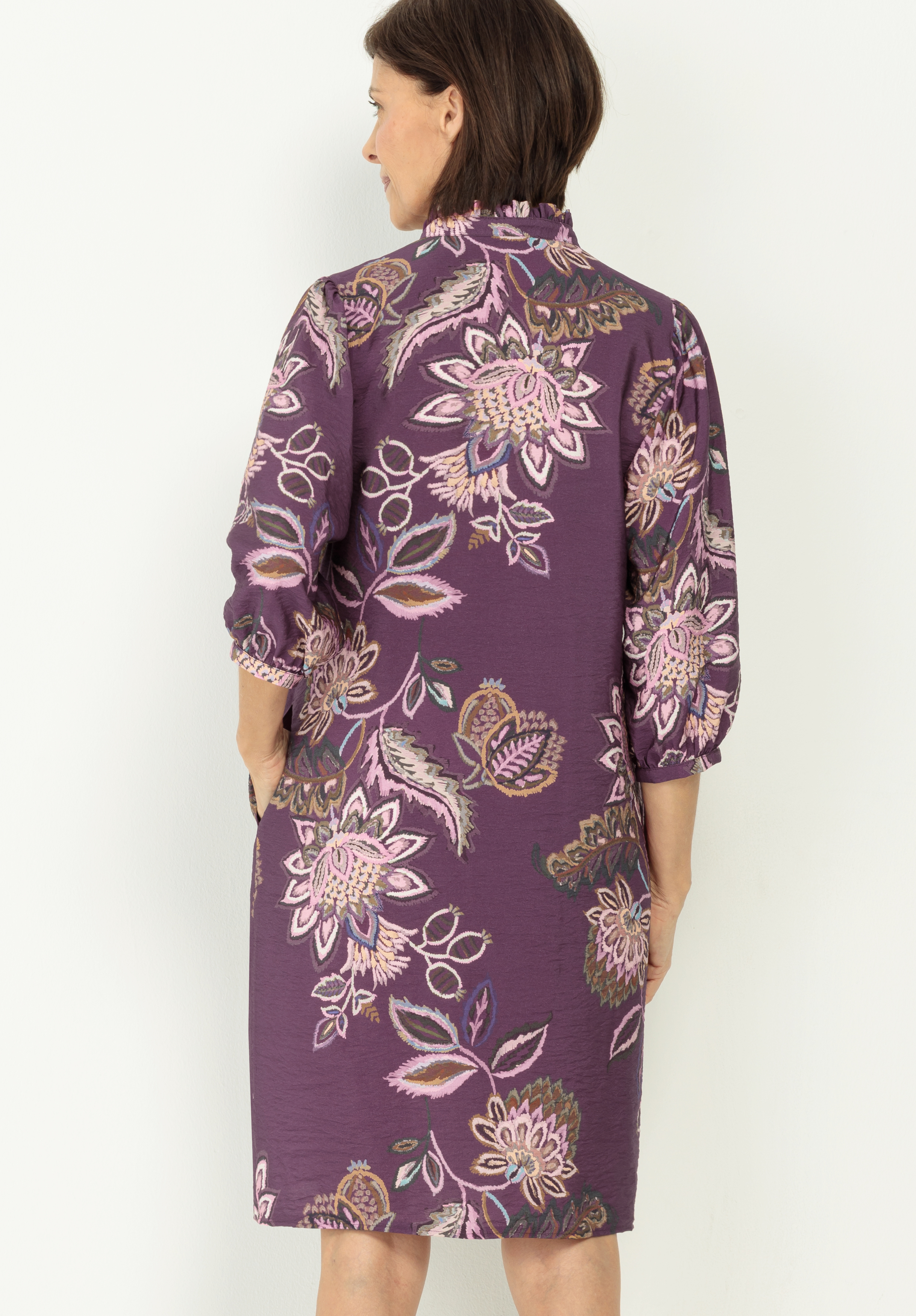 Eine Frau mit kurzen braunen Haaren steht mit dem Gesicht zur Wand und trägt ein lilafarbenes Kleid mit rosa und weißem Blumenmuster und Dreiviertelärmeln. Das Kleid fällt ihr bis zu den Knien und hat eine lockere, bequeme Passform.
