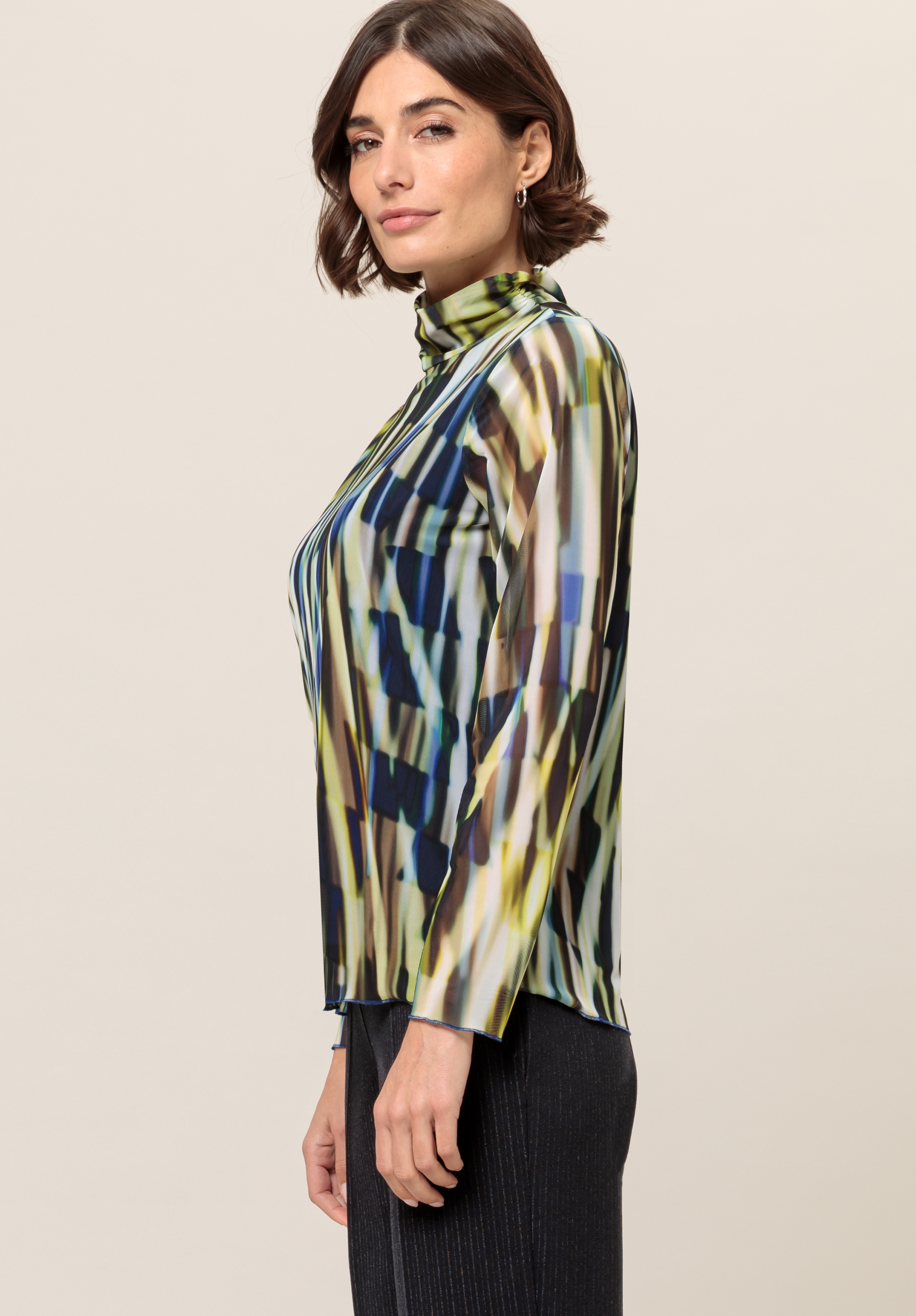 Frau mit kurzen braunen Haaren, die ein langärmliges, hochgeschlossenes Oberteil mit einem abstrakten geometrischen Muster in Blau, Grün, Gelb und Weiß trägt, dazu dunkle Hosen, und seitlich vor einem schlichten Hintergrund steht.