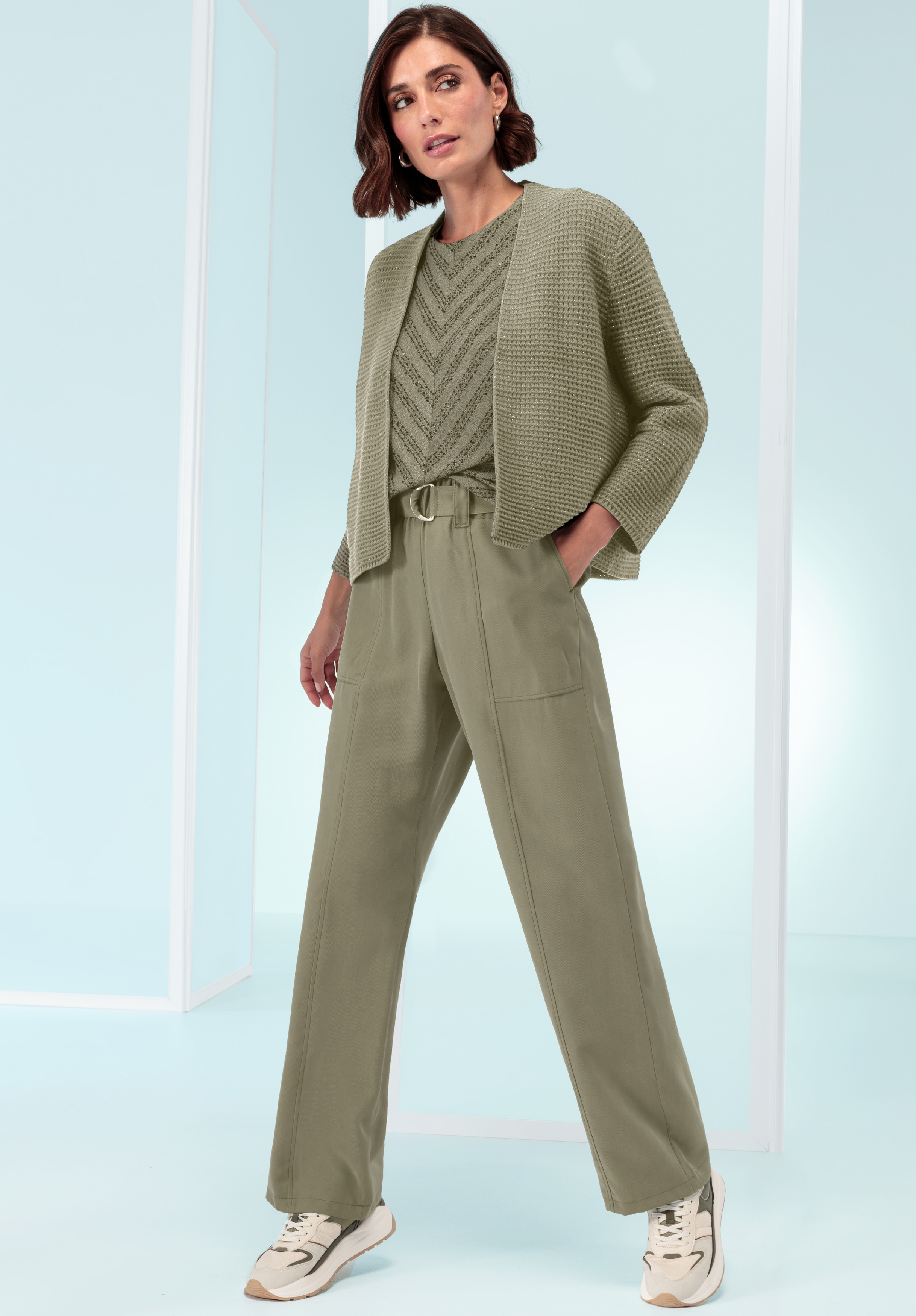 Eine Frau steht selbstbewusst vor einem hellblauen Hintergrund. Sie trägt eine salbeigrüne Hose mit weitem Bein, eine strukturierte Strickjacke, ein gemustertes Oberteil und weiße Turnschuhe.