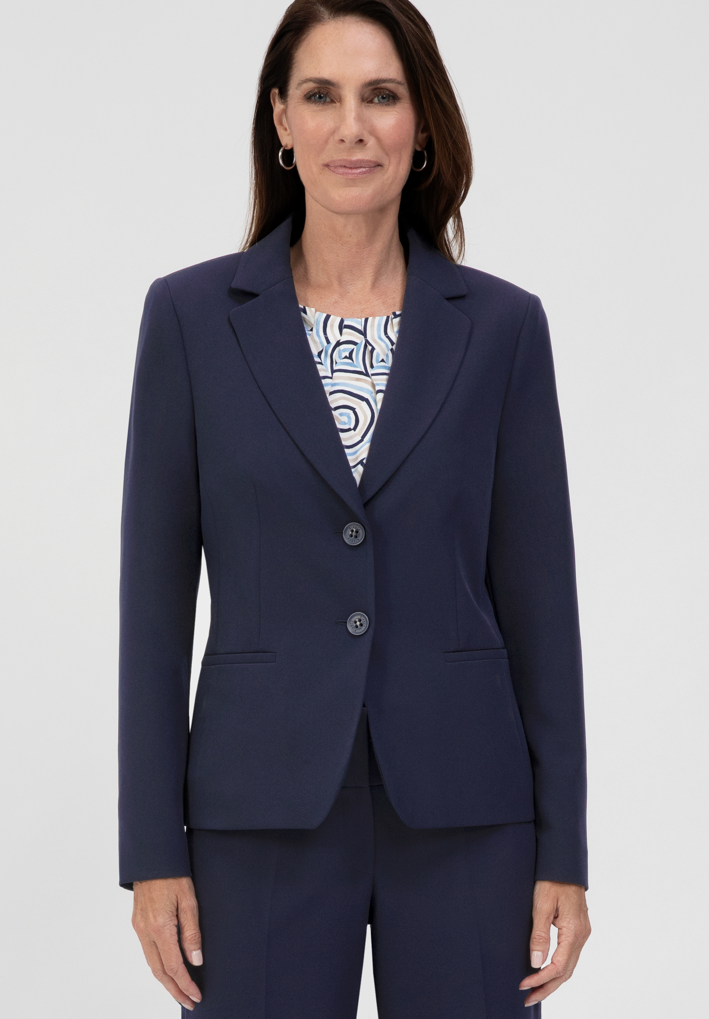 Eine Frau in einem marineblauen Blazer und einer dazu passenden Hose steht mit dem Blick nach vorne gerichtet da und lächelt leicht. Sie hat langes braunes Haar und trägt unter dem Blazer ein gemustertes Oberteil. Der Hintergrund ist einfarbig und hell.