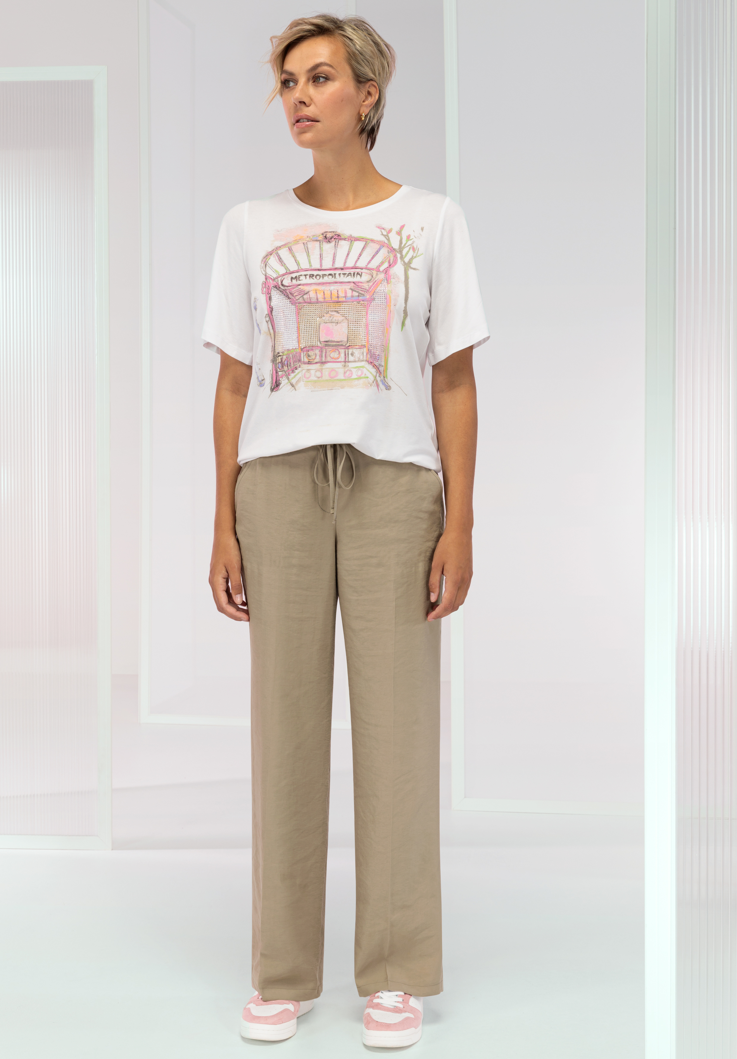 Eine Frau steht im Haus und trägt ein weißes T-Shirt mit buntem Aufdruck, weite beige Hosen und rosa-weiße Turnschuhe. Sie hat kurze, helle Haare und blickt mit neutralem Gesichtsausdruck leicht zur Seite.