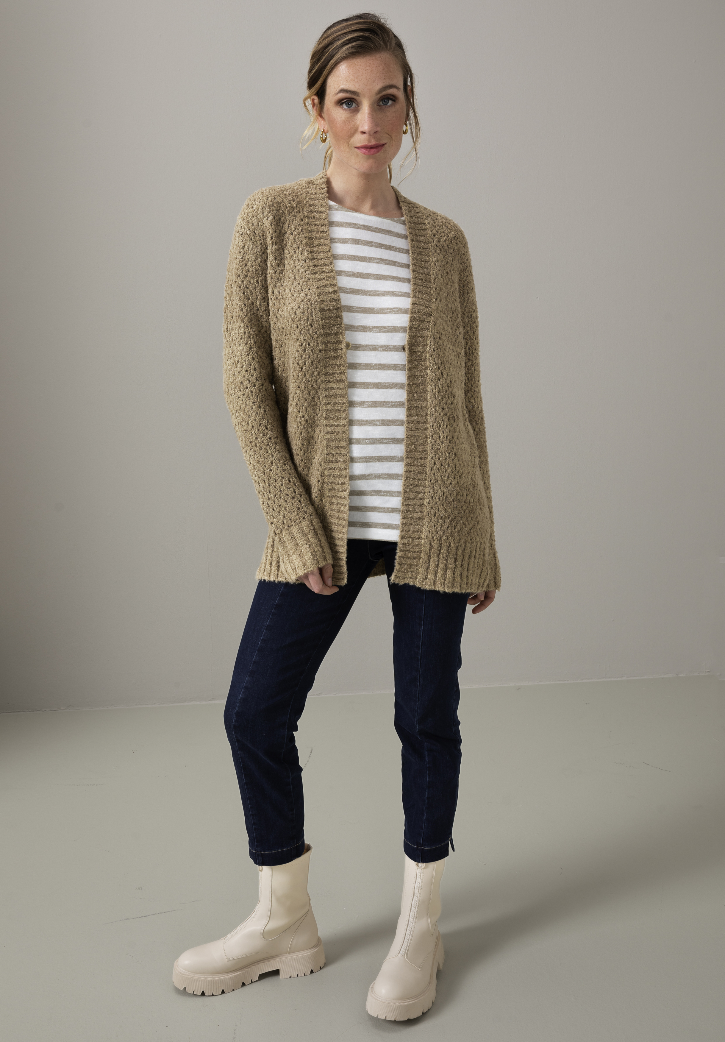 Eine Frau steht im Innenbereich und trägt eine beige Strickjacke über einem weiß-grau gestreiften Hemd, dunkelblaue Jeans und cremefarbene klobige Stiefel vor einem schlichten hellgrauen Hintergrund.