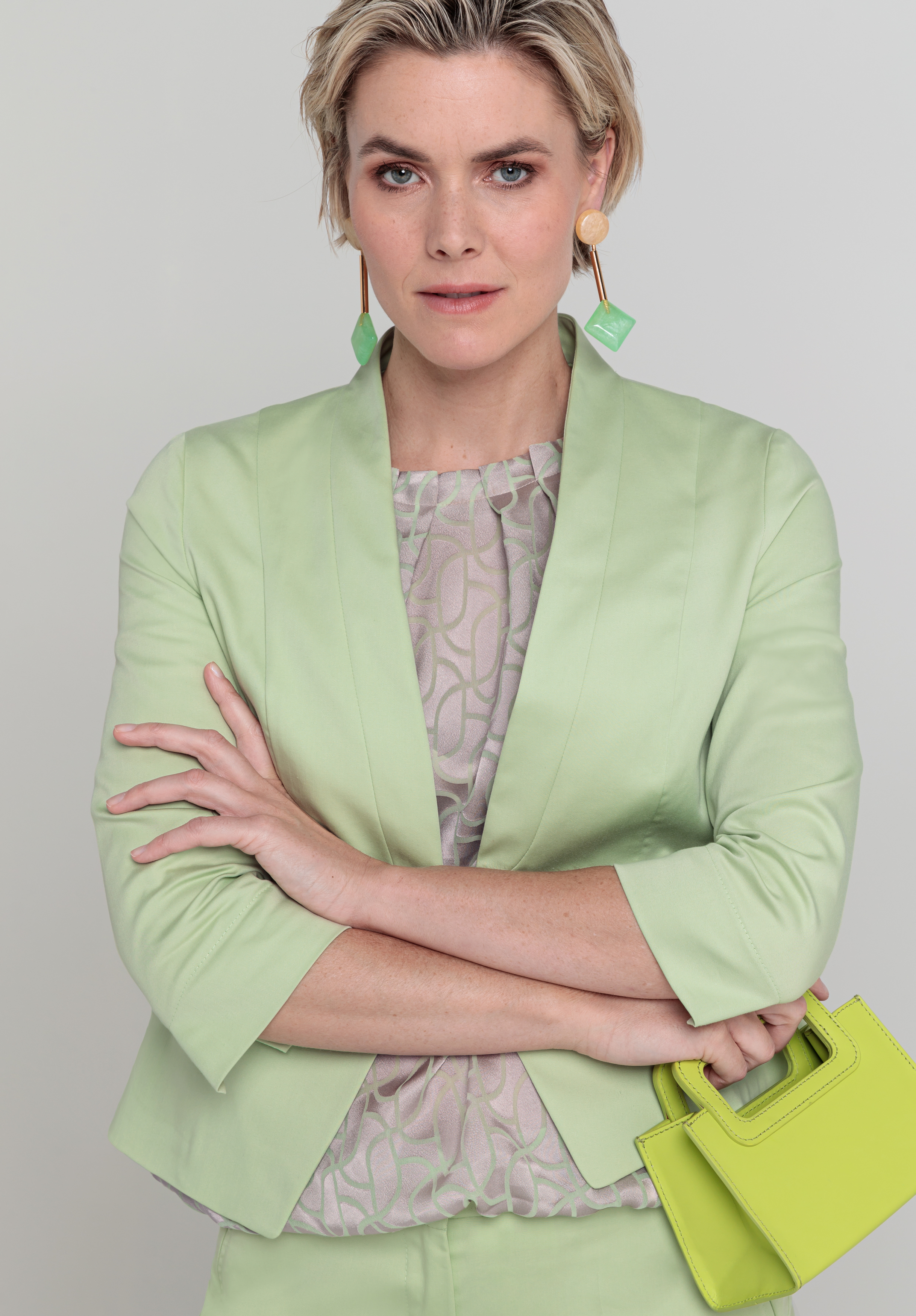 Eine Frau mit kurzen blonden Haaren trägt einen hellgrünen Blazer über einer gemusterten Bluse, goldene und grüne geometrische Ohrringe und hält eine kleine limettengrüne Handtasche in der Hand.