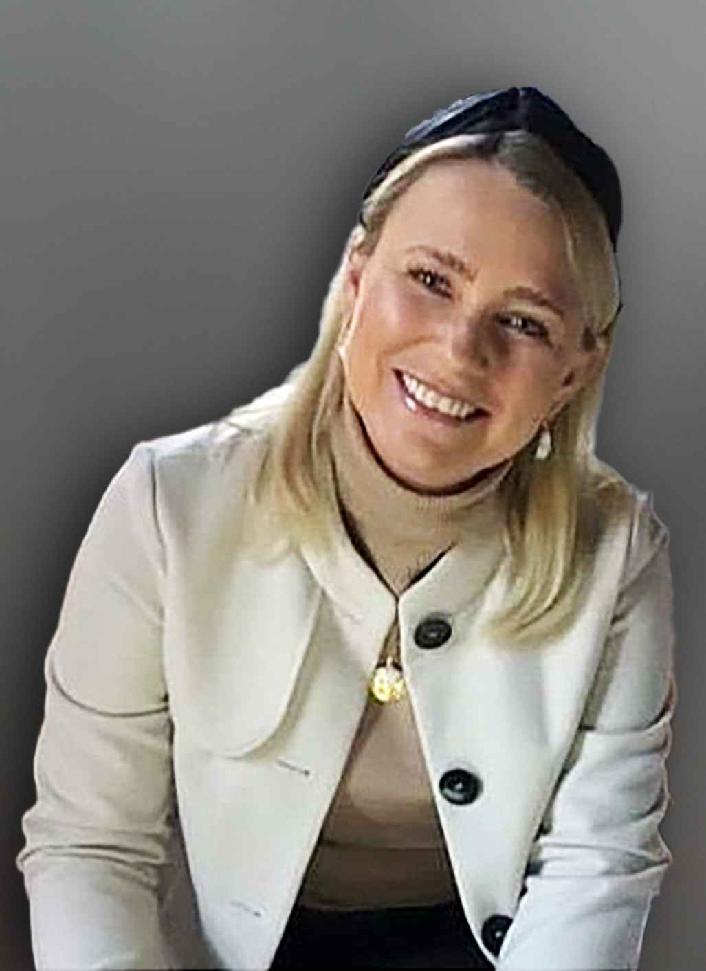 Eine Frau mit blondem Haar, die eine helle Jacke, einen beigen Rollkragenpullover und ein schwarzes Stirnband trägt, lächelt in die Kamera vor einem schlichten grauen Hintergrund.