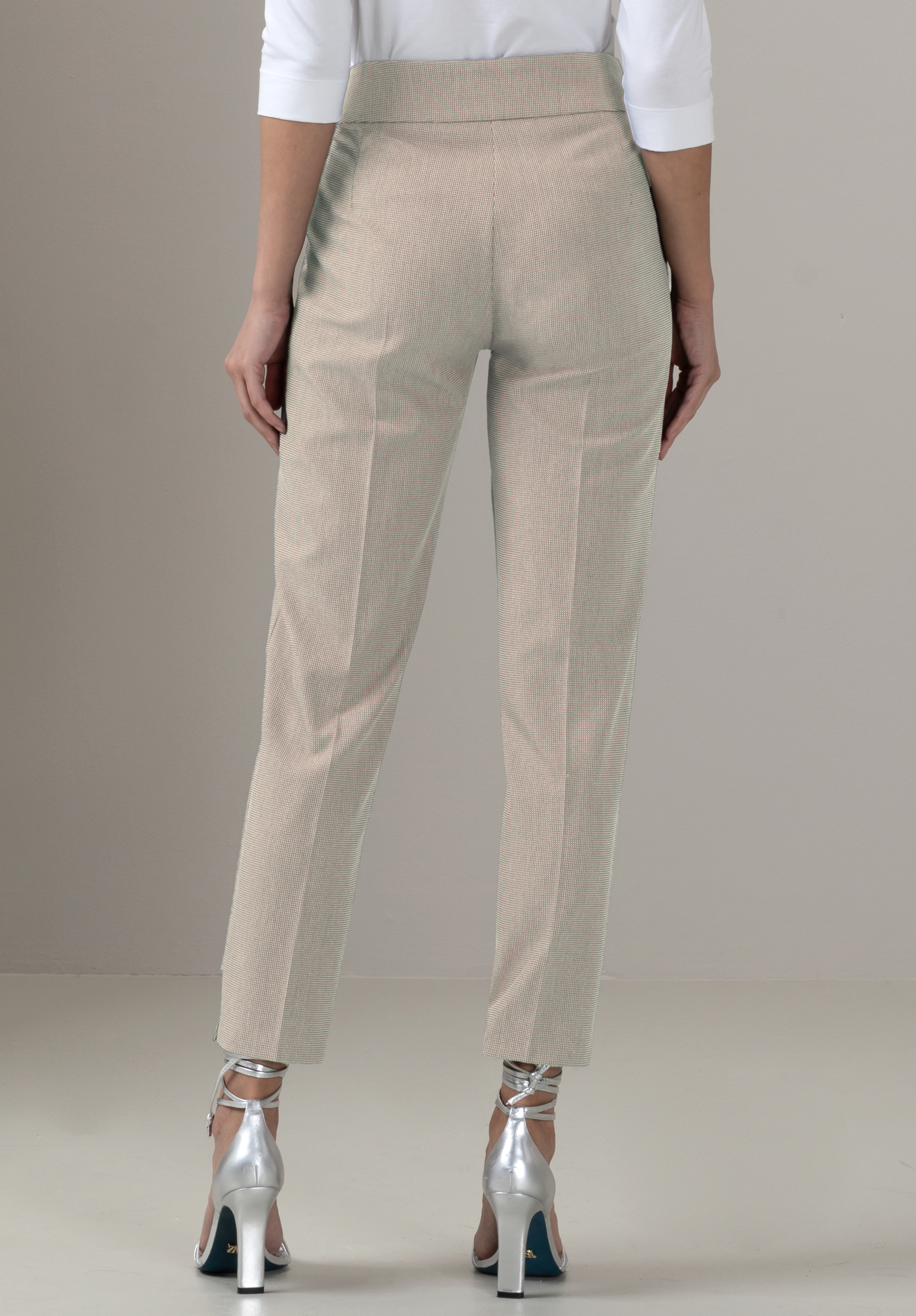 Eine Person, die eine hellbeige maßgeschneiderte Hose und ein weißes Oberteil trägt, wird von hinten gezeigt. Sie trägt außerdem metallisch-silberne hochhackige Sandalen mit Knöchelriemen und steht auf einem hellen Boden.