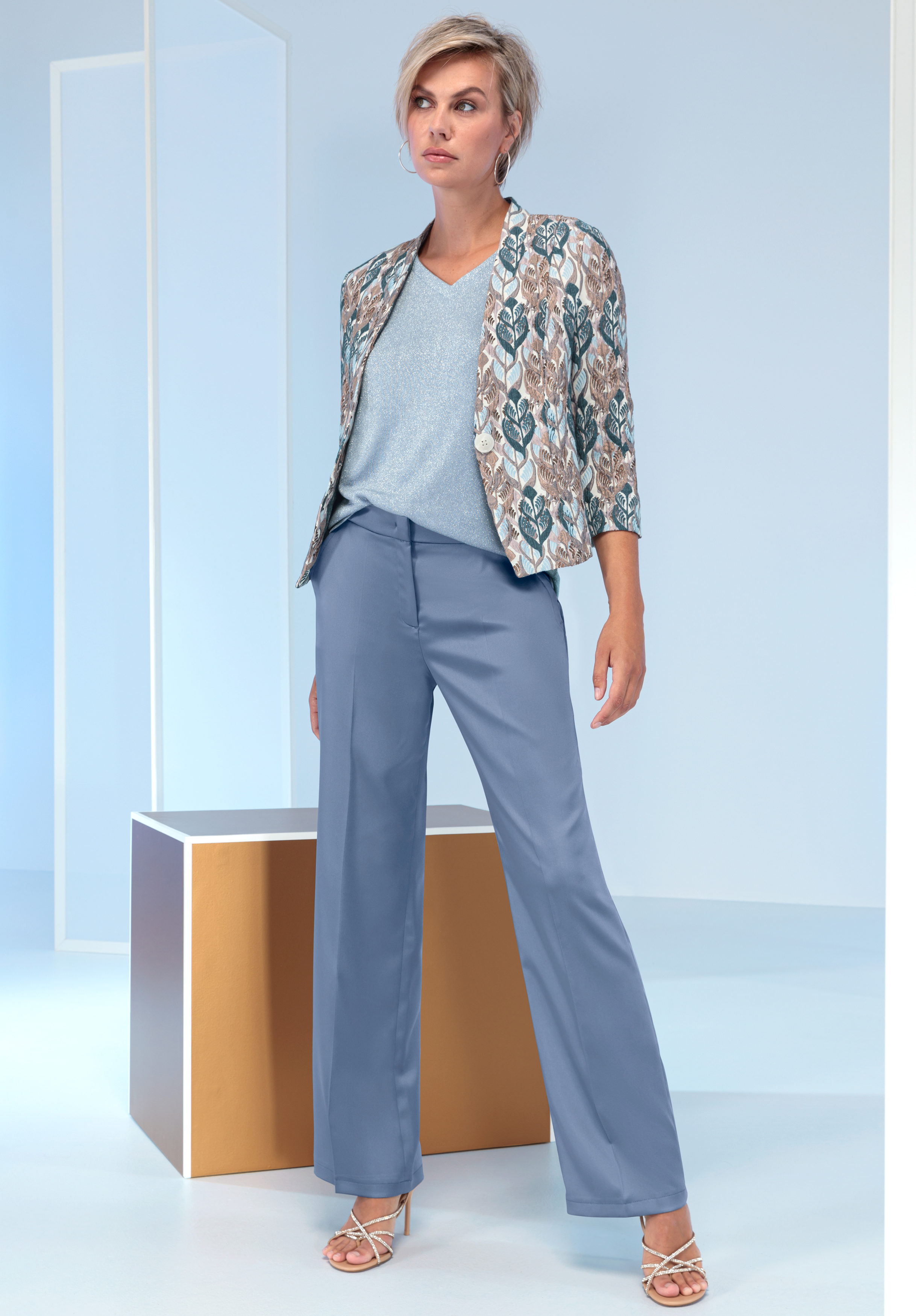 Eine Frau steht selbstbewusst in einer hellblauen Hose, einem hellblauen Oberteil, einer gemusterten Jacke in Blau- und Beigetönen und beigen Sandalen mit Absatz in einem modernen, minimalistischen Ambiente mit geometrischem Dekor.
