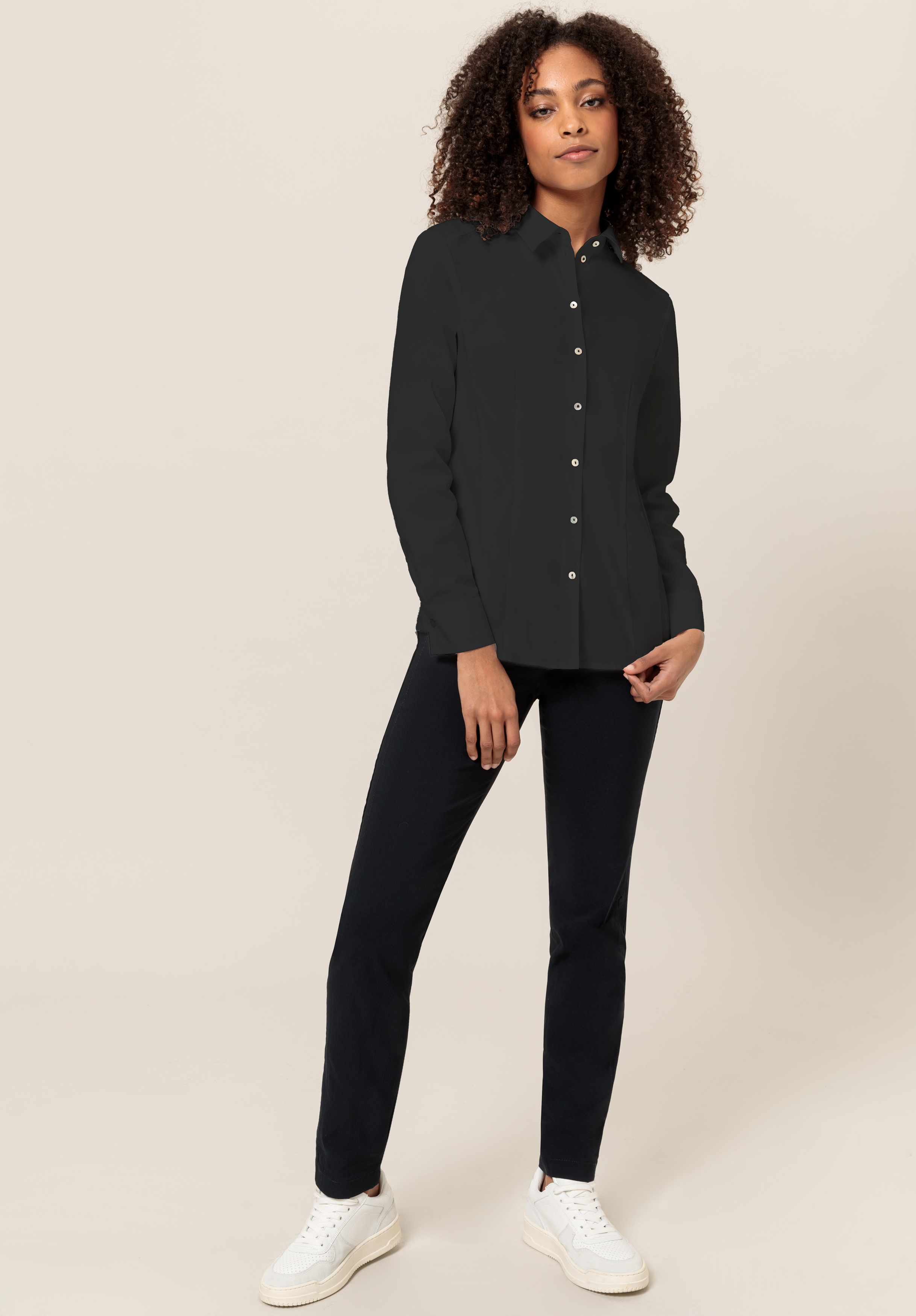 Eine Frau mit lockigem Haar steht vor einem hellen Hintergrund und trägt ein schwarzes Button-up-Hemd, eine schwarze Hose und weiße Turnschuhe. Sie blickt mit entspannter Miene in die Kamera und hat eine Hand an der Seite.