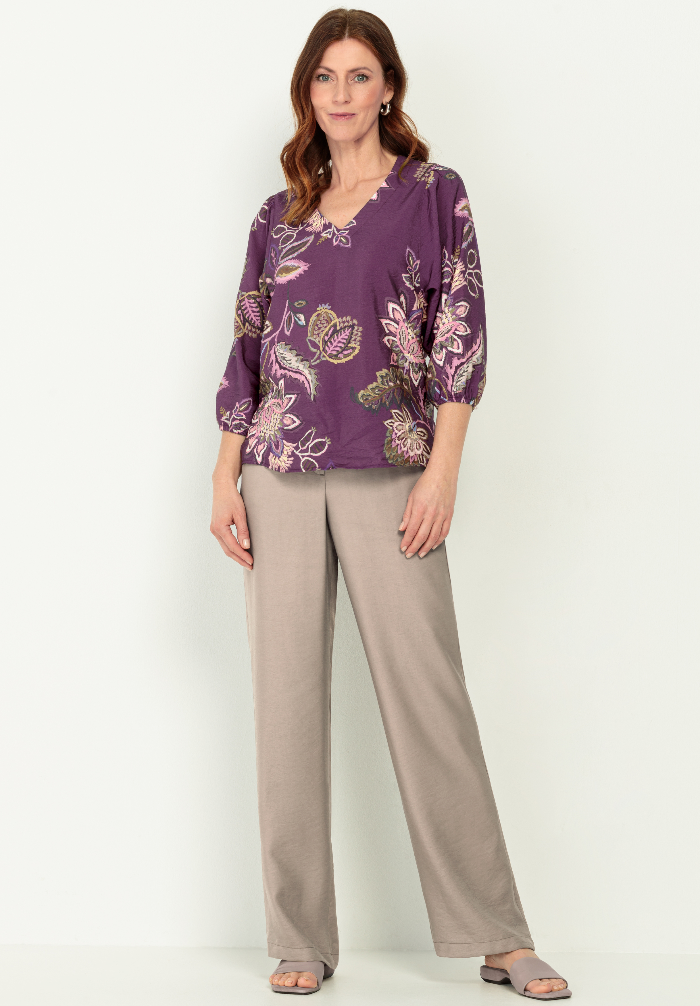 Eine Frau steht vor einem hellen Hintergrund und trägt eine lila geblümte Bluse mit Dreiviertelärmeln, eine beigefarbene Hose mit weitem Bein und helle Sandalen. Sie hat gewelltes braunes Haar und einen entspannten, freundlichen Gesichtsausdruck.