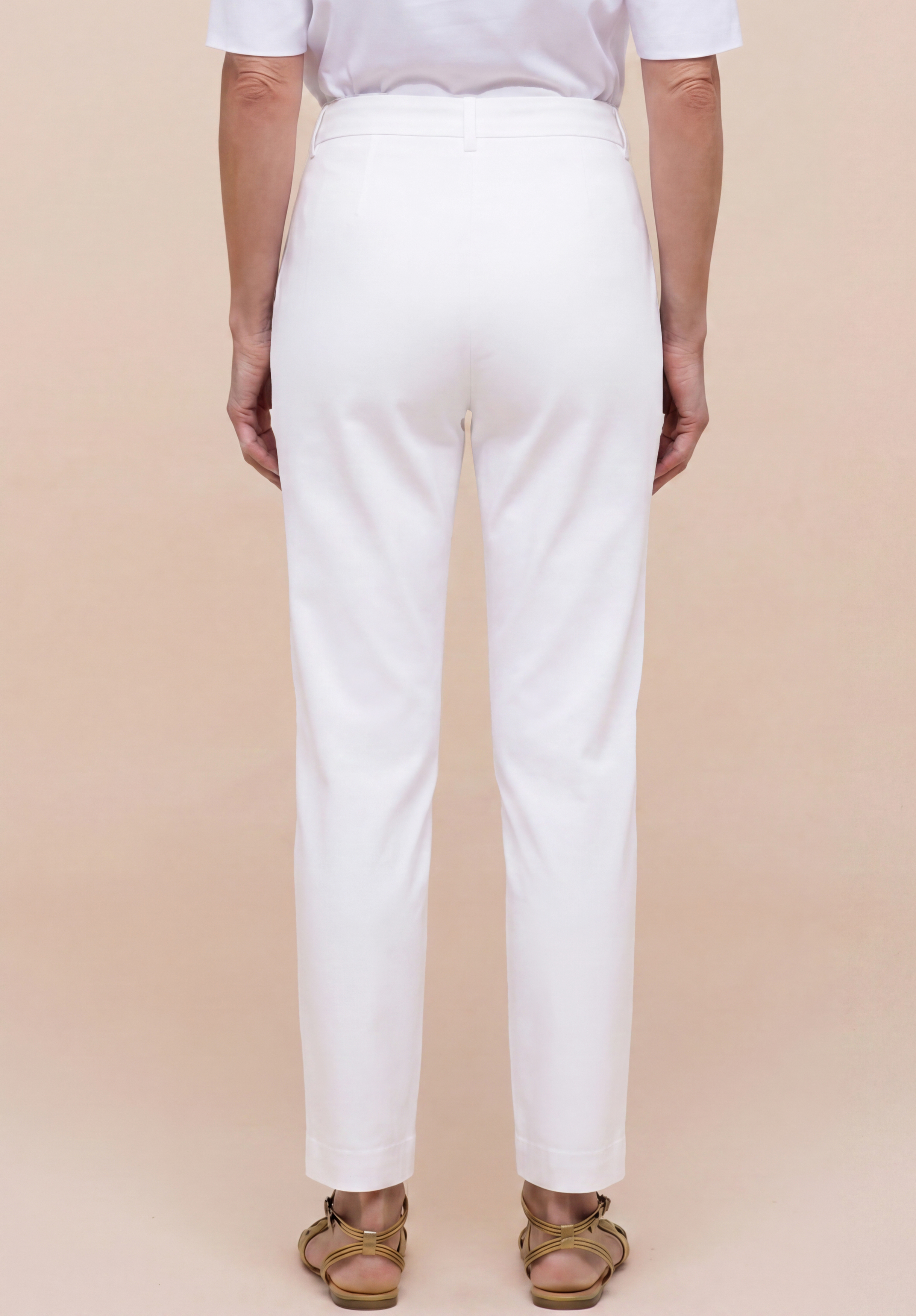 Eine Person steht mit dem Rücken zur Kamera und trägt eine weiße Hose mit hoher Taille, ein weißes Kurzarmhemd sowie flache, hellbraune Sandalen. Der Hintergrund ist einfarbig beige.