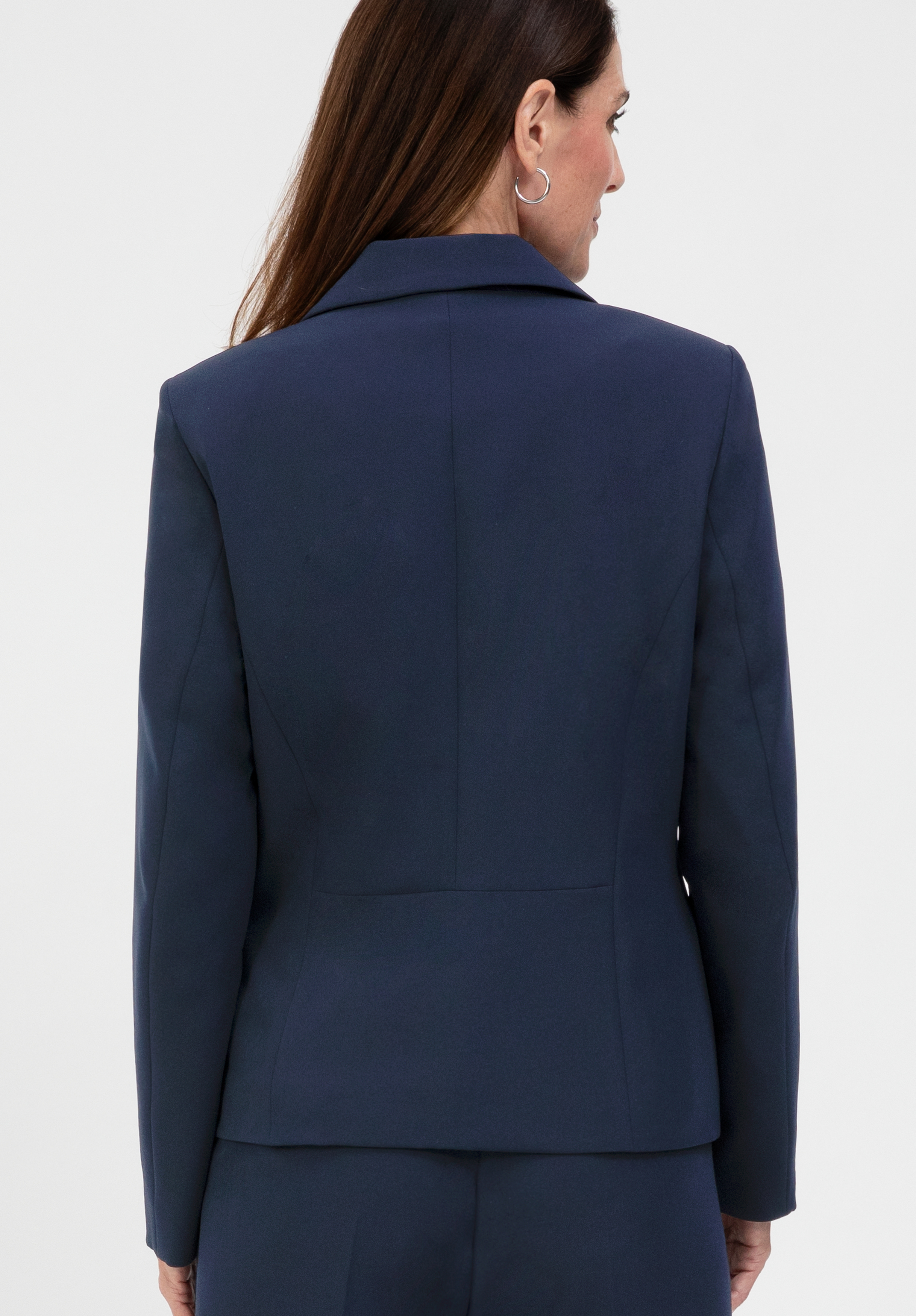 Eine Frau mit langen braunen Haaren ist von hinten zu sehen; sie trägt einen marineblauen, taillierten Blazer und eine dazu passende Hose vor einem schlichten weißen Hintergrund.