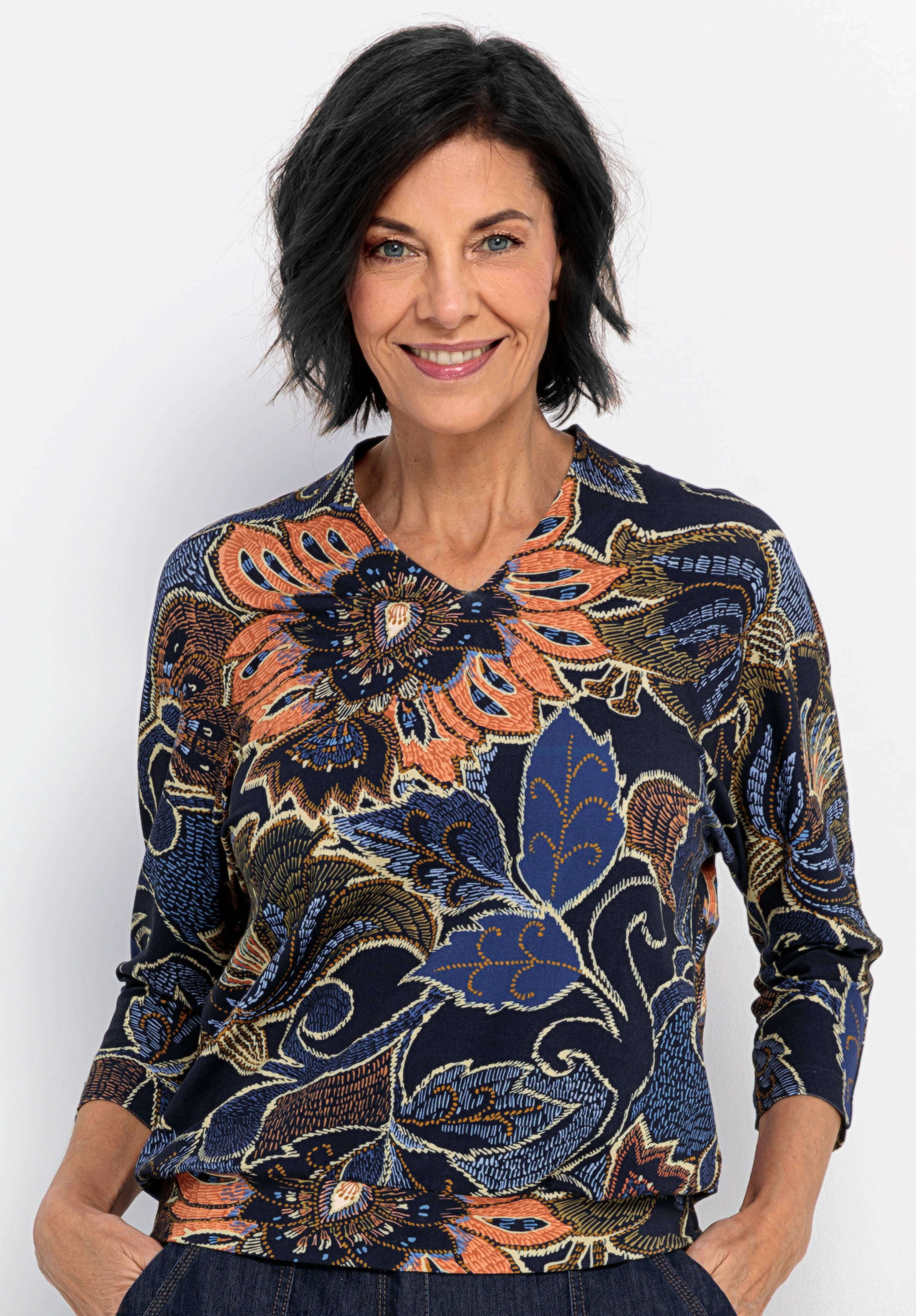 Eine Frau mit kurzen schwarzen Haaren, die ein langärmeliges Hemd mit einem auffälligen Blumenmuster in Blau, Orange und Beige trägt, steht vor einem schlichten hellen Hintergrund und lächelt, die Hände in den Taschen.