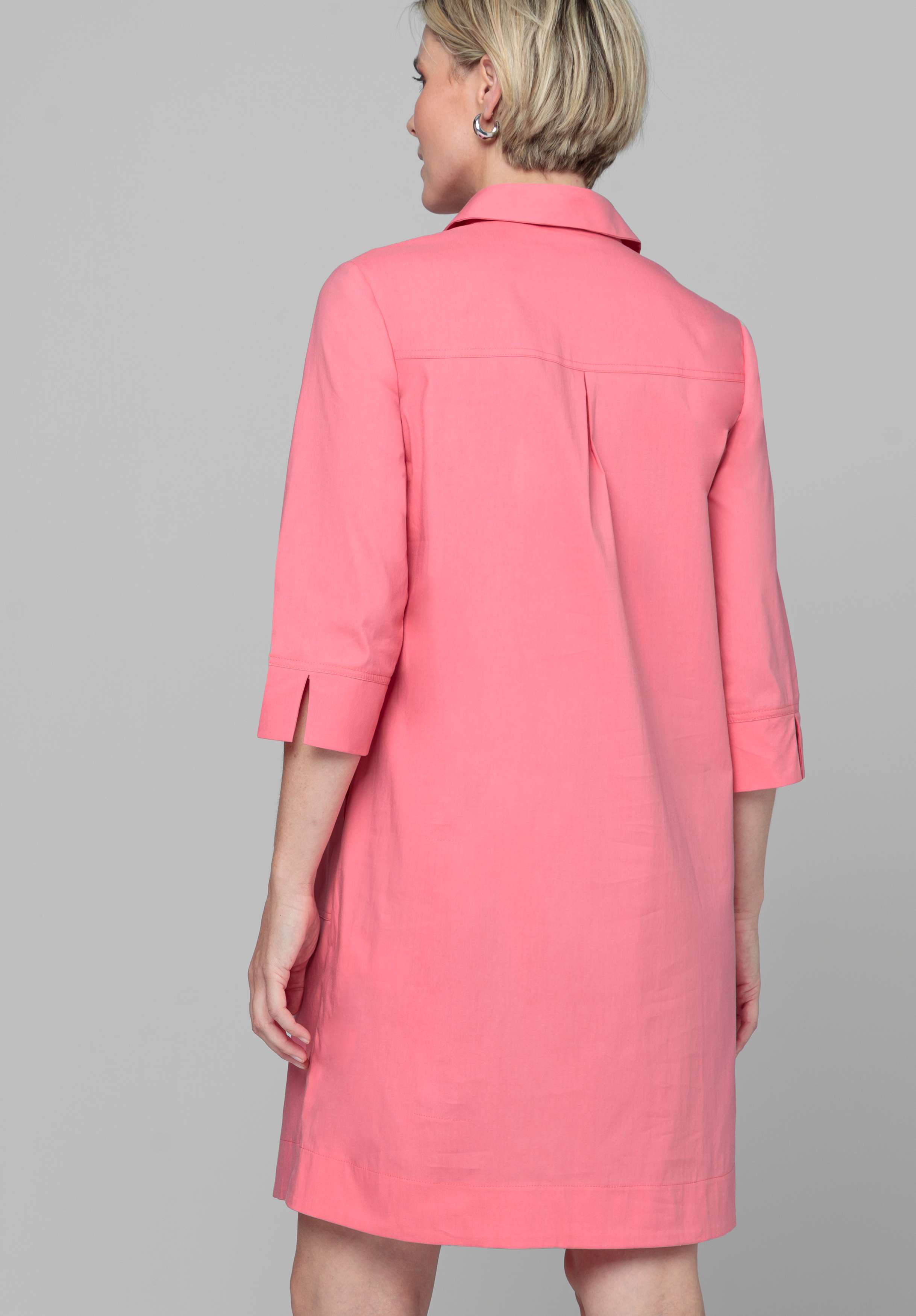 Von hinten ist eine Person mit kurzen blonden Haaren zu sehen, die ein knielanges, korallenrosa Kleid mit Dreiviertelärmeln und Kragen trägt und vor einem schlichten grauen Hintergrund steht.