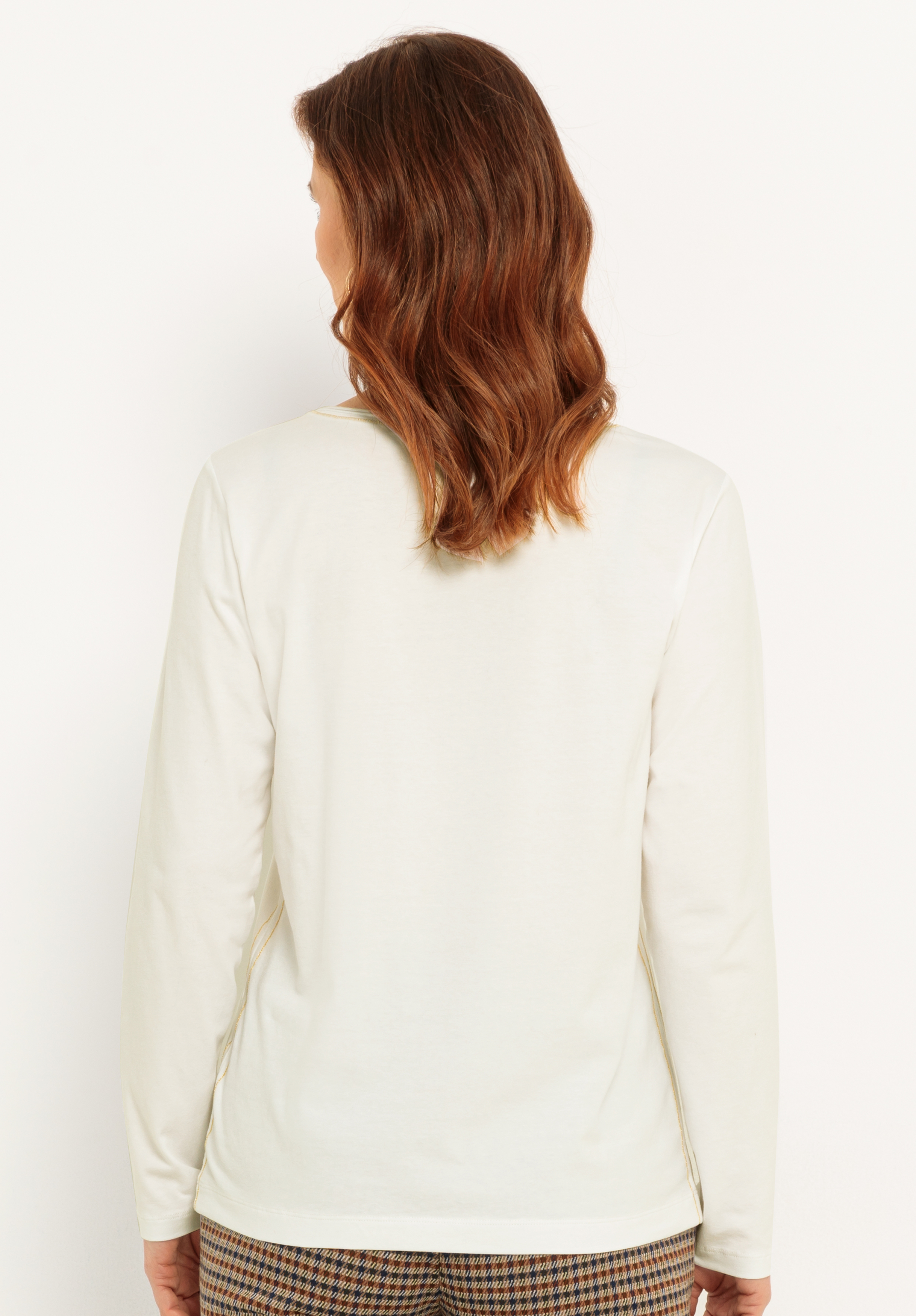 Eine Frau mit schulterlangen braunen Haaren wird von hinten gezeigt. Sie trägt ein langärmeliges, cremefarbenes Oberteil und eine karierte Hose. Sie steht vor einem schlichten weißen Hintergrund.