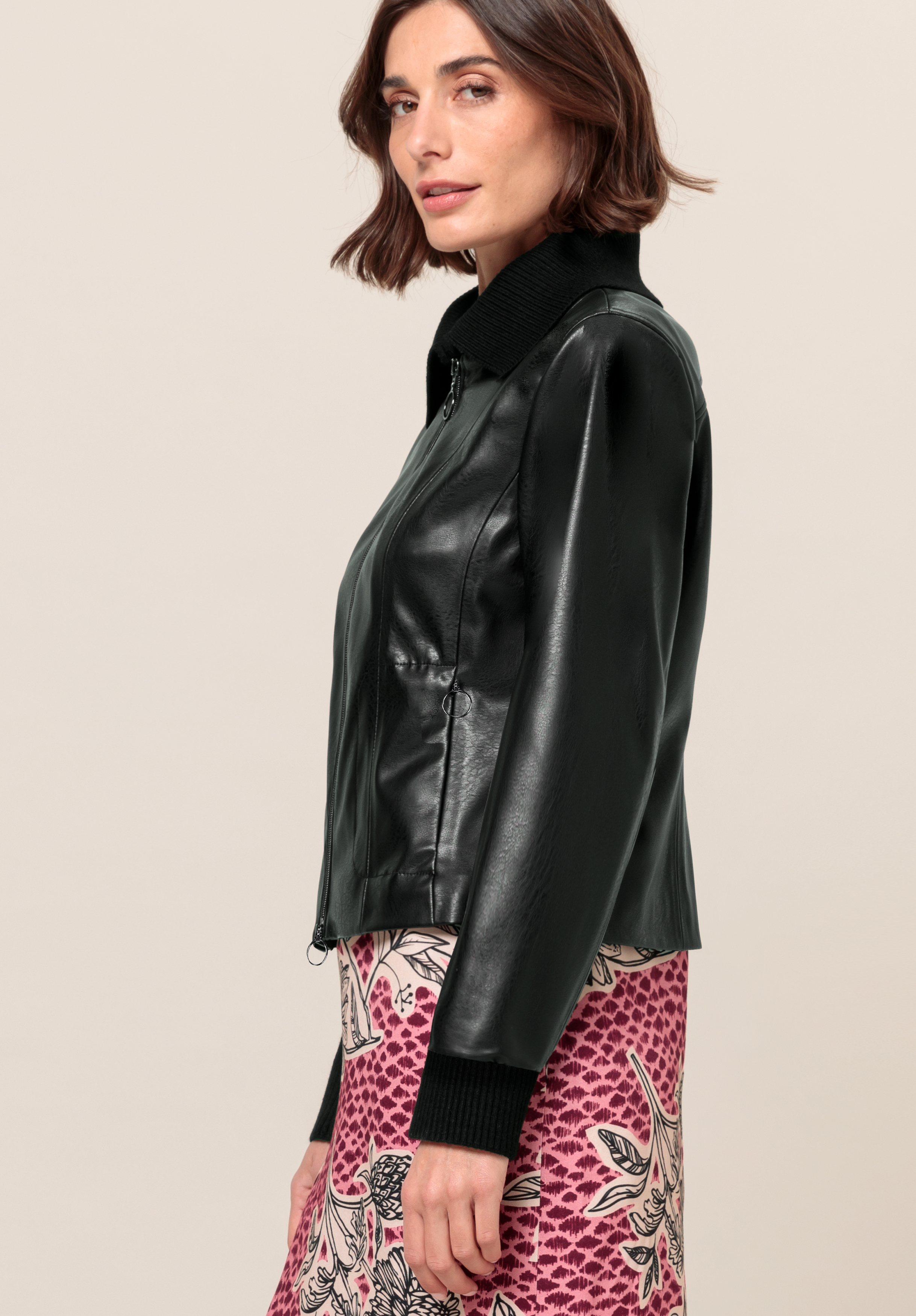 Eine Frau mit schulterlangem braunem Haar trägt eine schwarze Lederjacke mit gerippten Bündchen und Kragen sowie einen rosa-weiß gemusterten Rock. Sie steht seitlich und blickt in die Kamera vor einem schlichten Hintergrund.