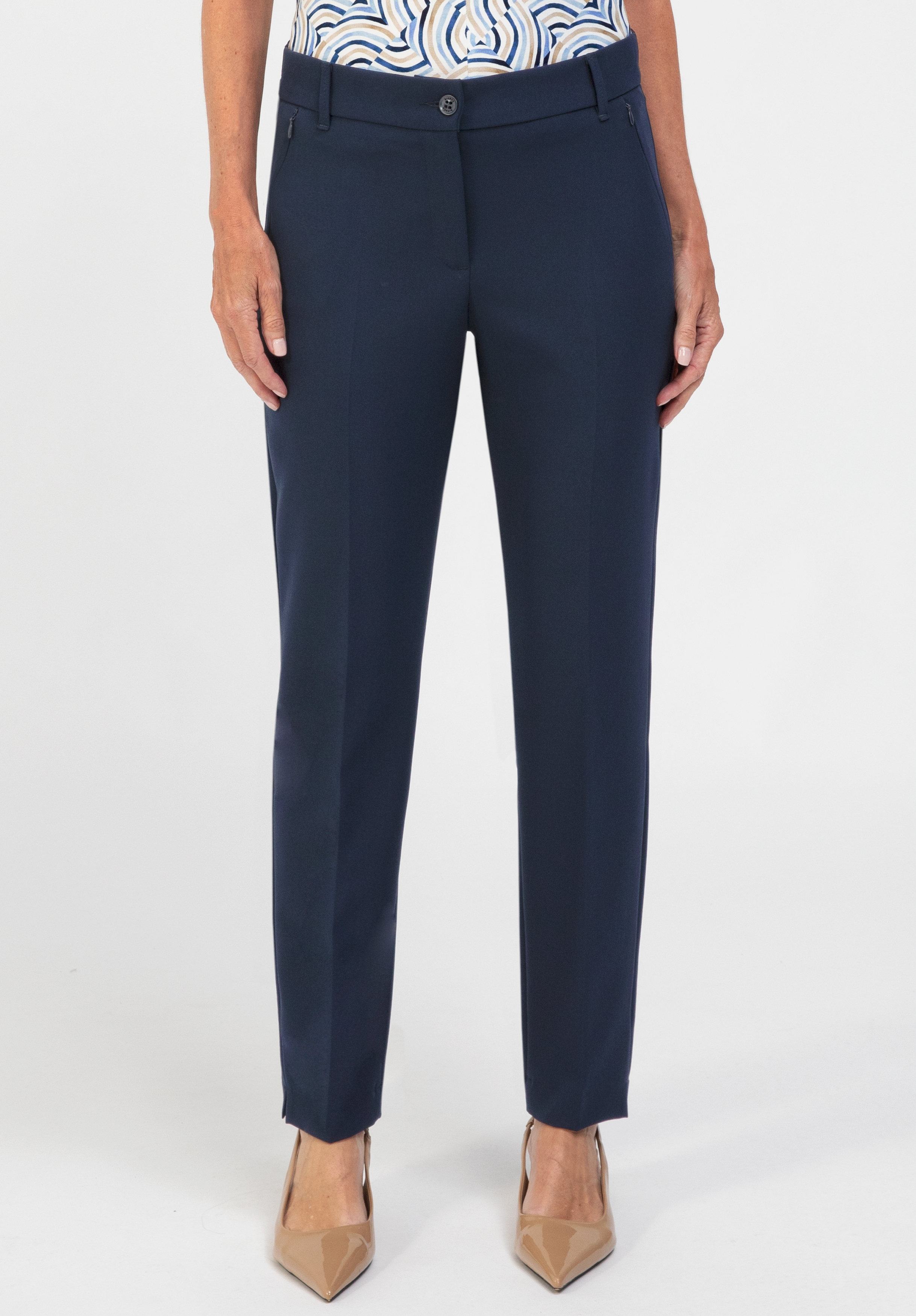 Eine Person, die eine marineblaue, maßgeschneiderte Hose und beige Pumps mit spitzer Zehenpartie trägt und vor einem schlichten weißen Hintergrund steht. Die Hose hat vorne einen Knopf- und Reißverschluss und wird mit einem gemusterten Oberteil kombiniert