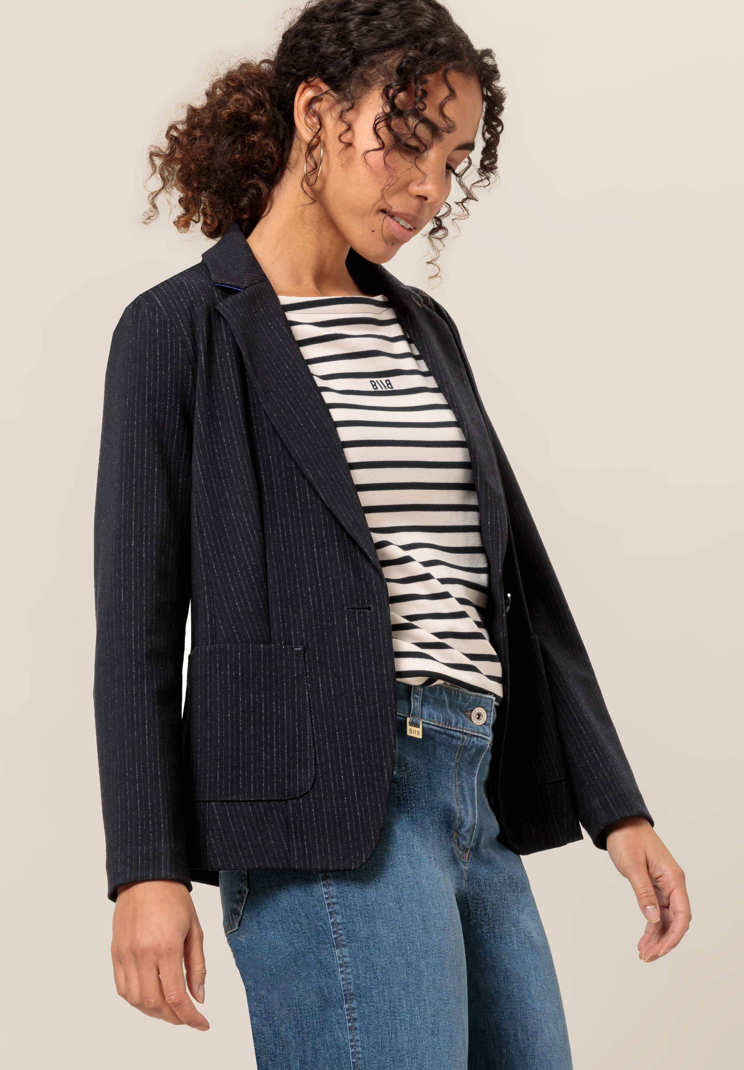 Eine Person mit lockigem Haar trägt einen schwarzen Nadelstreifenblazer über einem schwarz-weiß gestreiften Hemd mit „Oui“-Schriftzug, dazu Bluejeans und steht vor einem schlichten hellen Hintergrund.