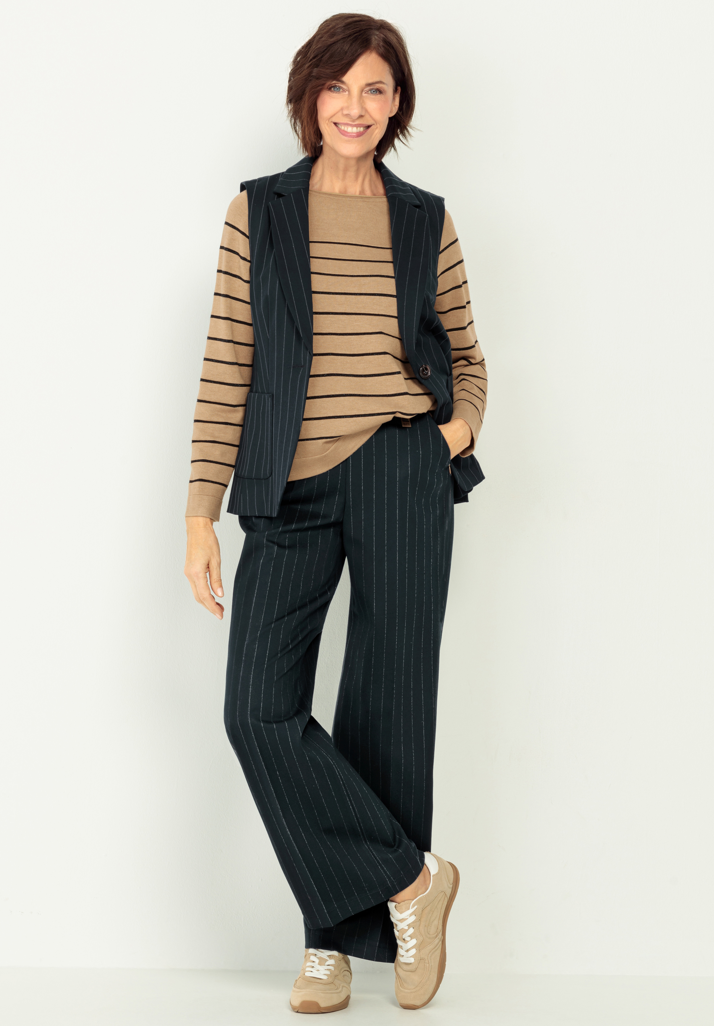 Eine Frau trägt einen hellbraunen und schwarz gestreiften Pullover, eine schwarze Nadelstreifenweste und eine passende Hose sowie beigefarbene Turnschuhe. Eine Hand in ihrer Tasche.