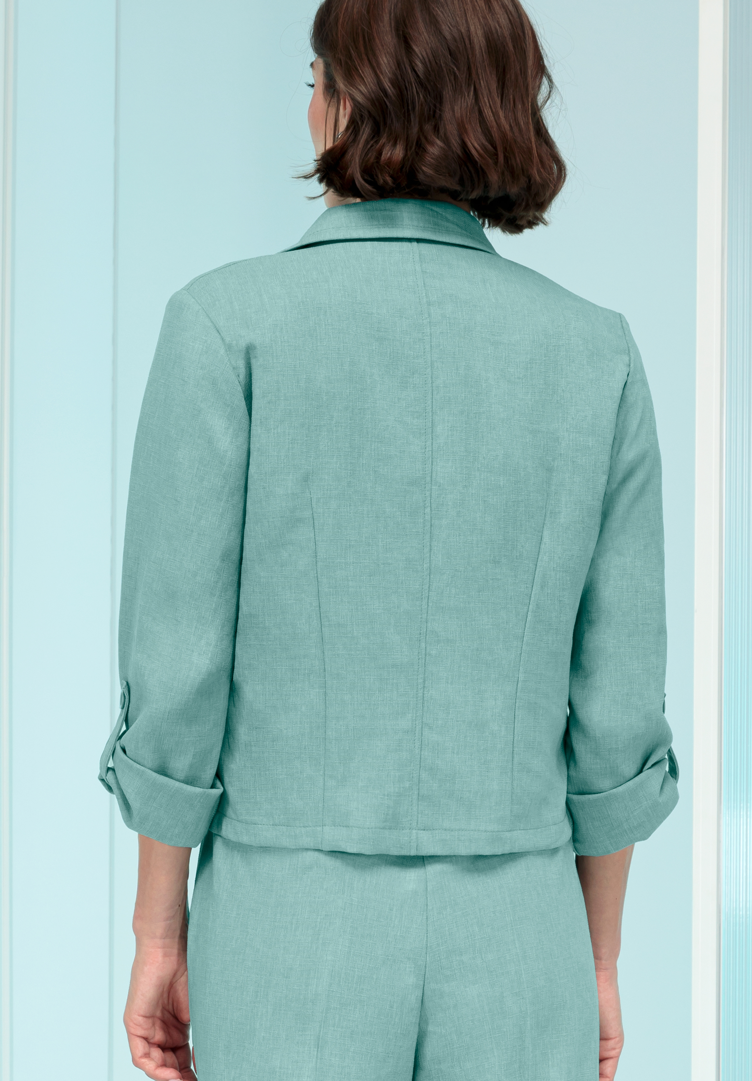Von hinten ist eine Person mit kurzen braunen Haaren zu sehen, die einen hellblauen Blazer mit hochgekrempelten Ärmeln und eine passende Hose trägt und vor einem hellblauen Hintergrund steht.