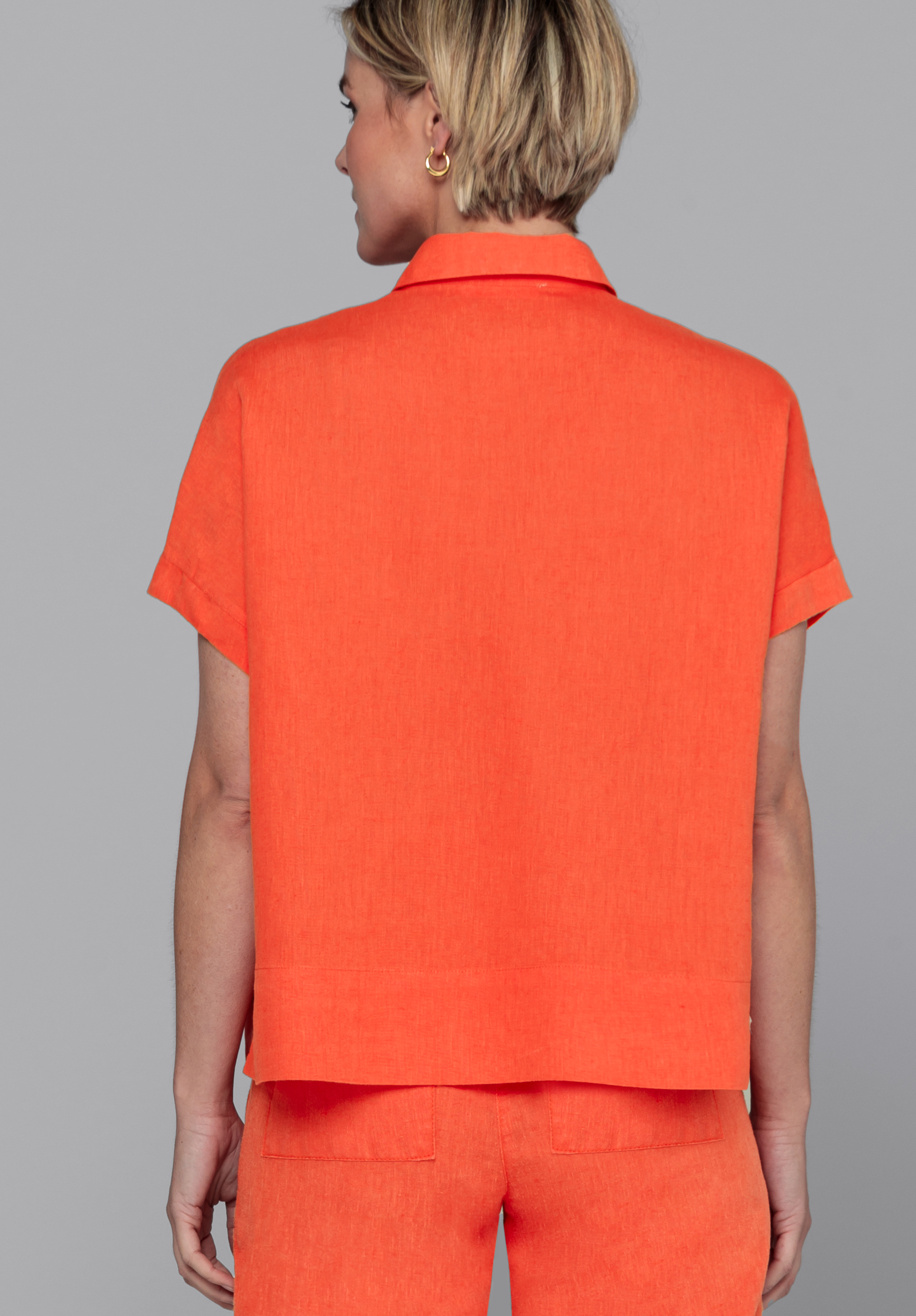 Eine Person mit kurzen blonden Haaren, die ein orangefarbenes kurzärmeliges Hemd mit Kragen und dazu passende orangefarbene Shorts trägt, steht mit dem Rücken zur Kamera vor einem schlichten grauen Hintergrund.