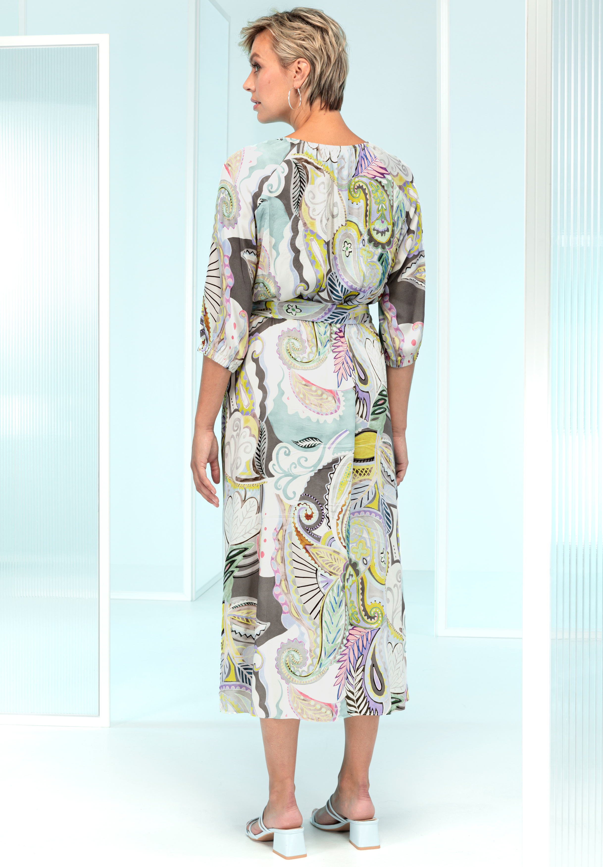 Eine Frau mit kurzen blonden Haaren steht in einem hellen, modernen Raum. Sie trägt ein langes, transparentes, gemustertes Kleid mit pastellfarbenen Wirbeln und Paisleymustern. Rückseite des Kleides und weiße Sandalen sind zu sehen.
